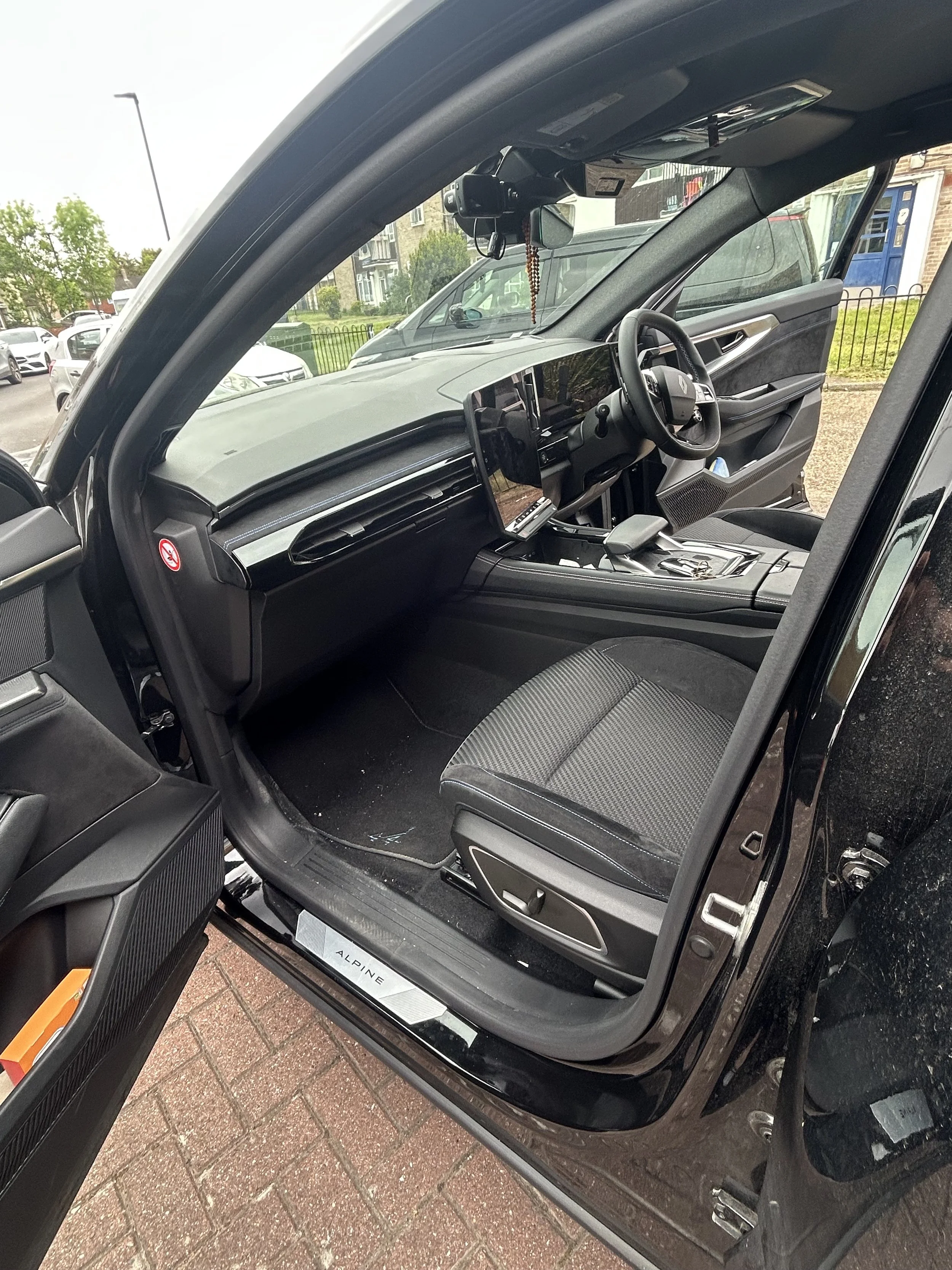 The interior of a black car showing the driver's side door open, with a black fabric seat, steering wheel, dashboard, and parking lot with other vehicles and buildings outside.