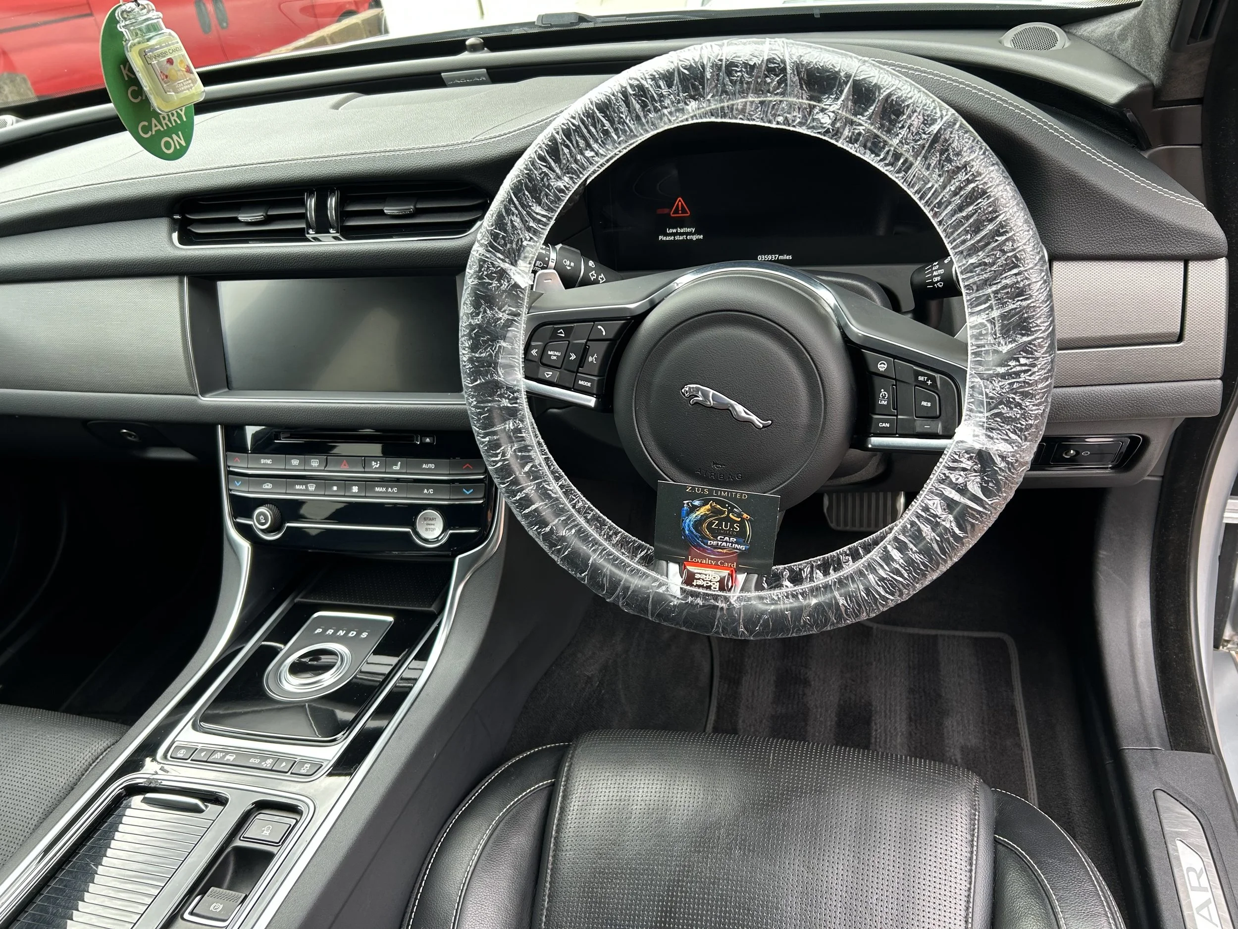The interior of a car showing the dashboard, steering wheel wrapped in plastic, and center console. The dashboard displays a warning message about a low battery. An air freshener hangs from the rearview mirror.