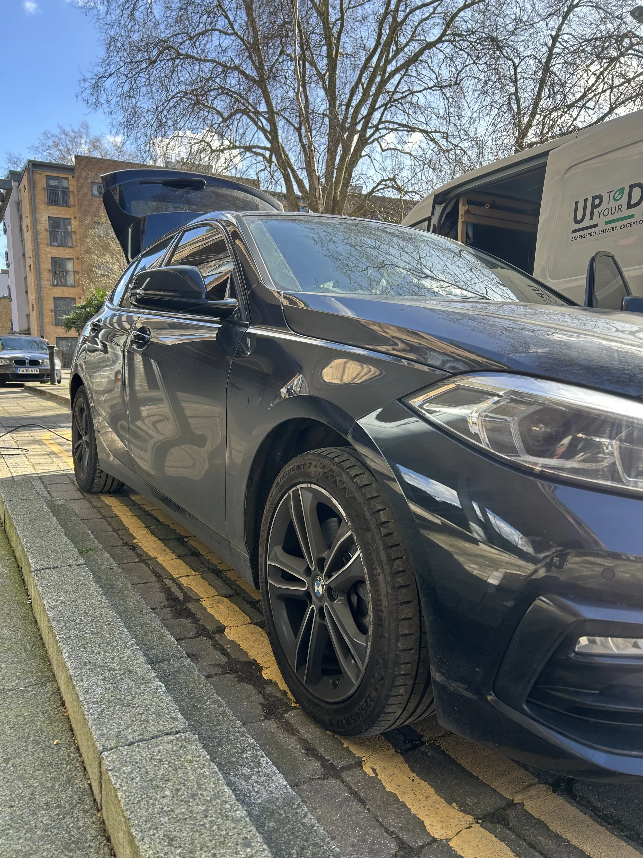 A silver BMW car parked in a parking lot with a large tree in the background and a delivery van nearby.