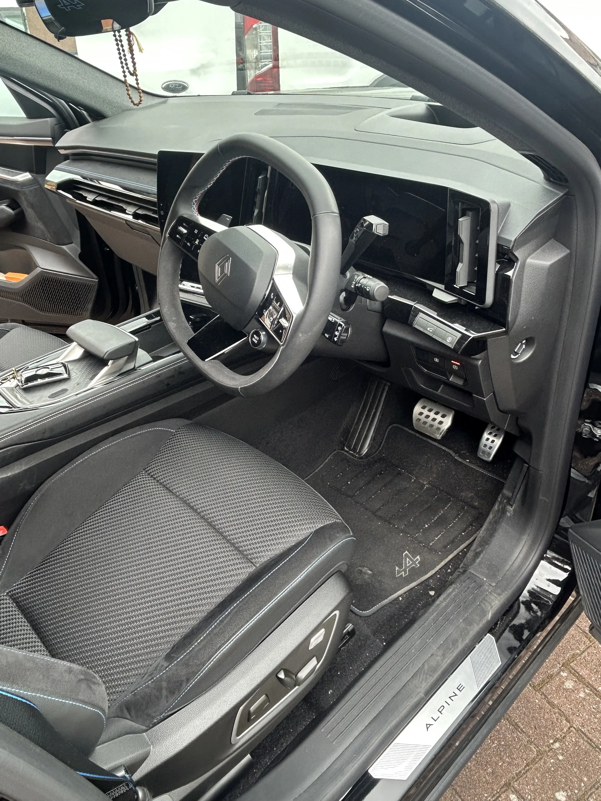 The interior of a modern vehicle featuring a steering wheel with vehicle controls, a digital dashboard, a mounted smartphone holder, and a seat with textured fabric. The floor has sporty metal pedals, and a logo on the door sill reads 'ALPINE'.
