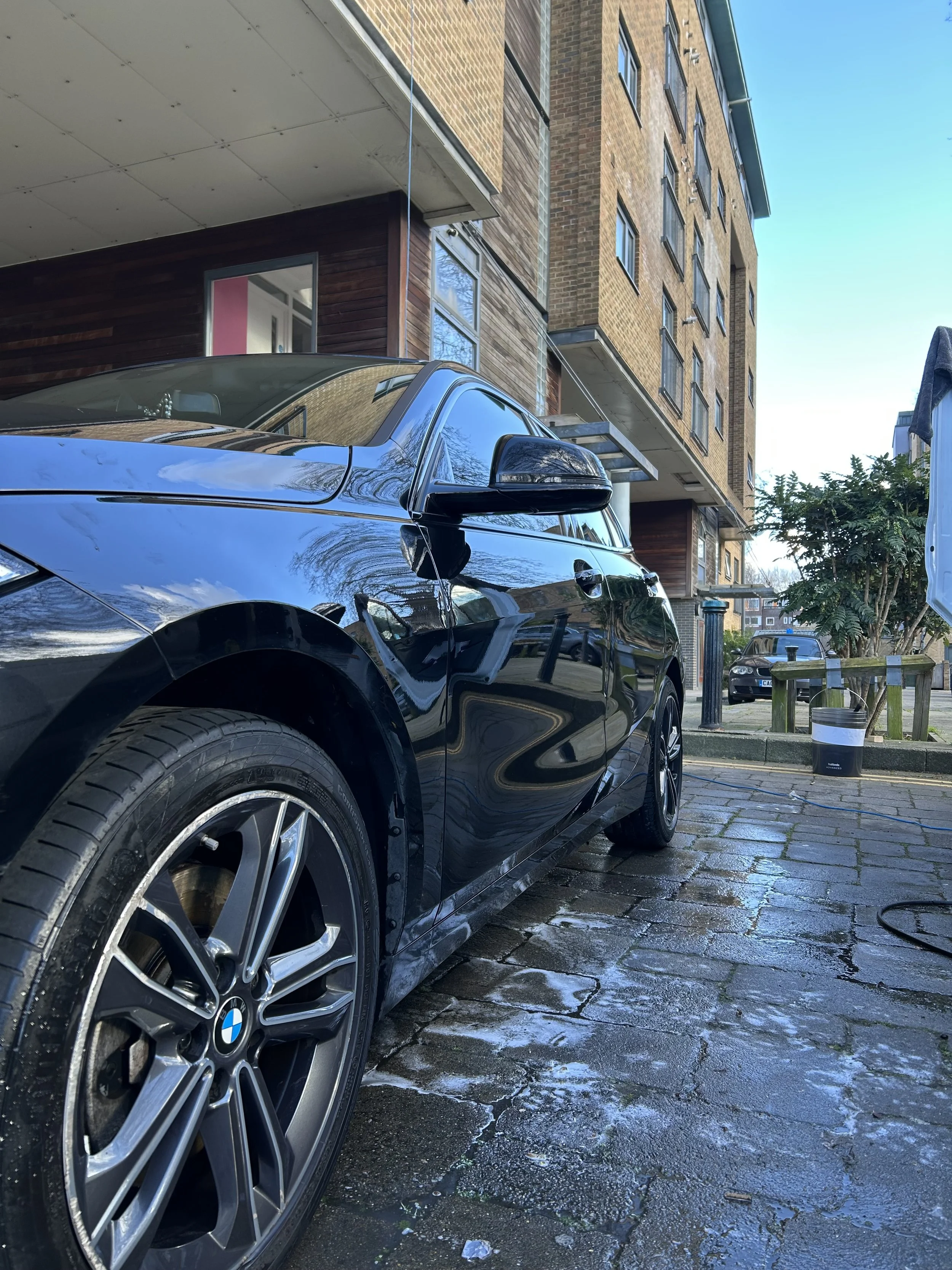 Black BMW car parked on a wet cobblestone street with modern brick apartment buildings in the background, blue sky overhead, and some greenery.