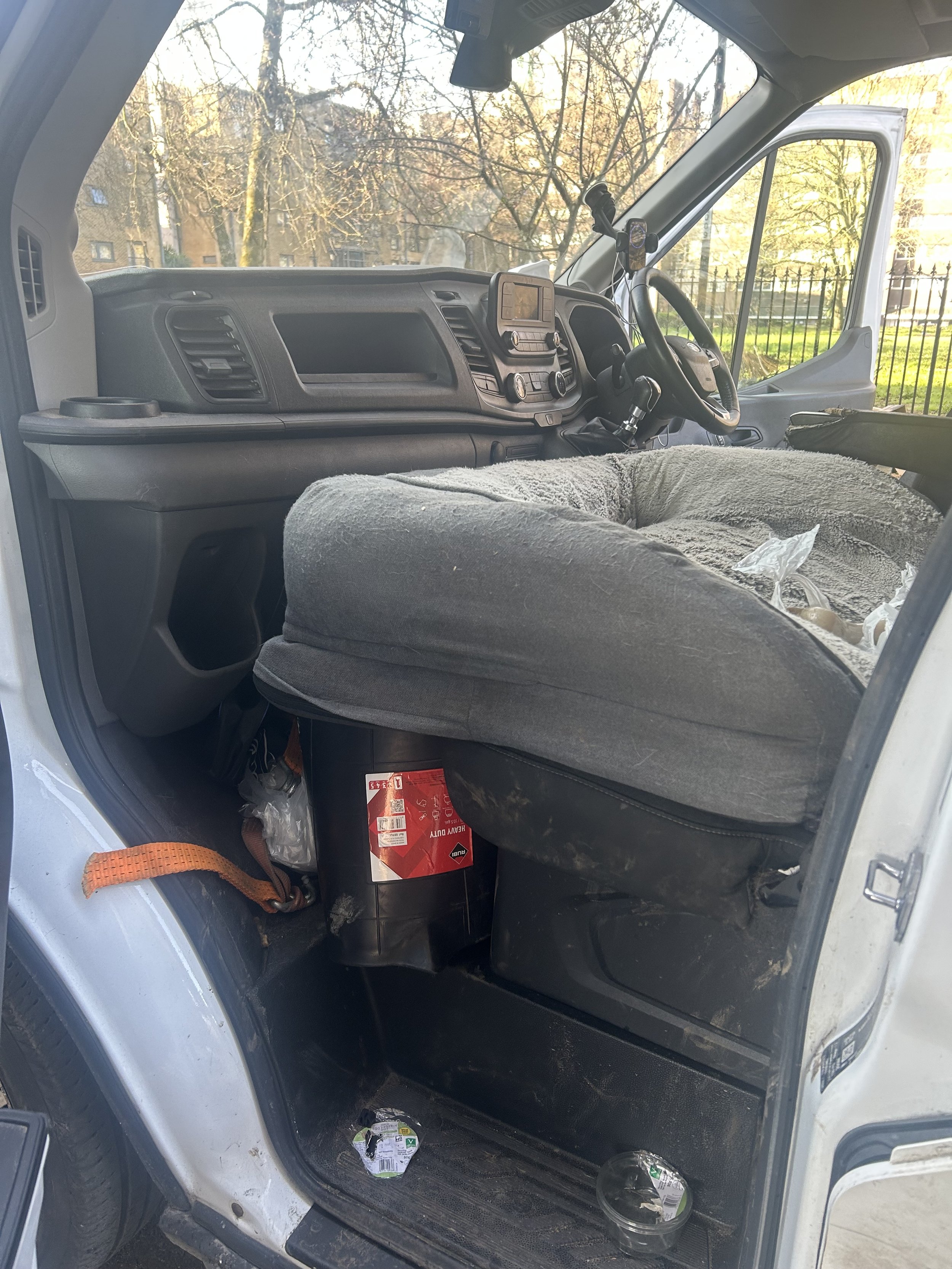 View inside a vehicle's cab showing a steering wheel, dashboard, and front seat. Outside trees are visible through the windshield.
