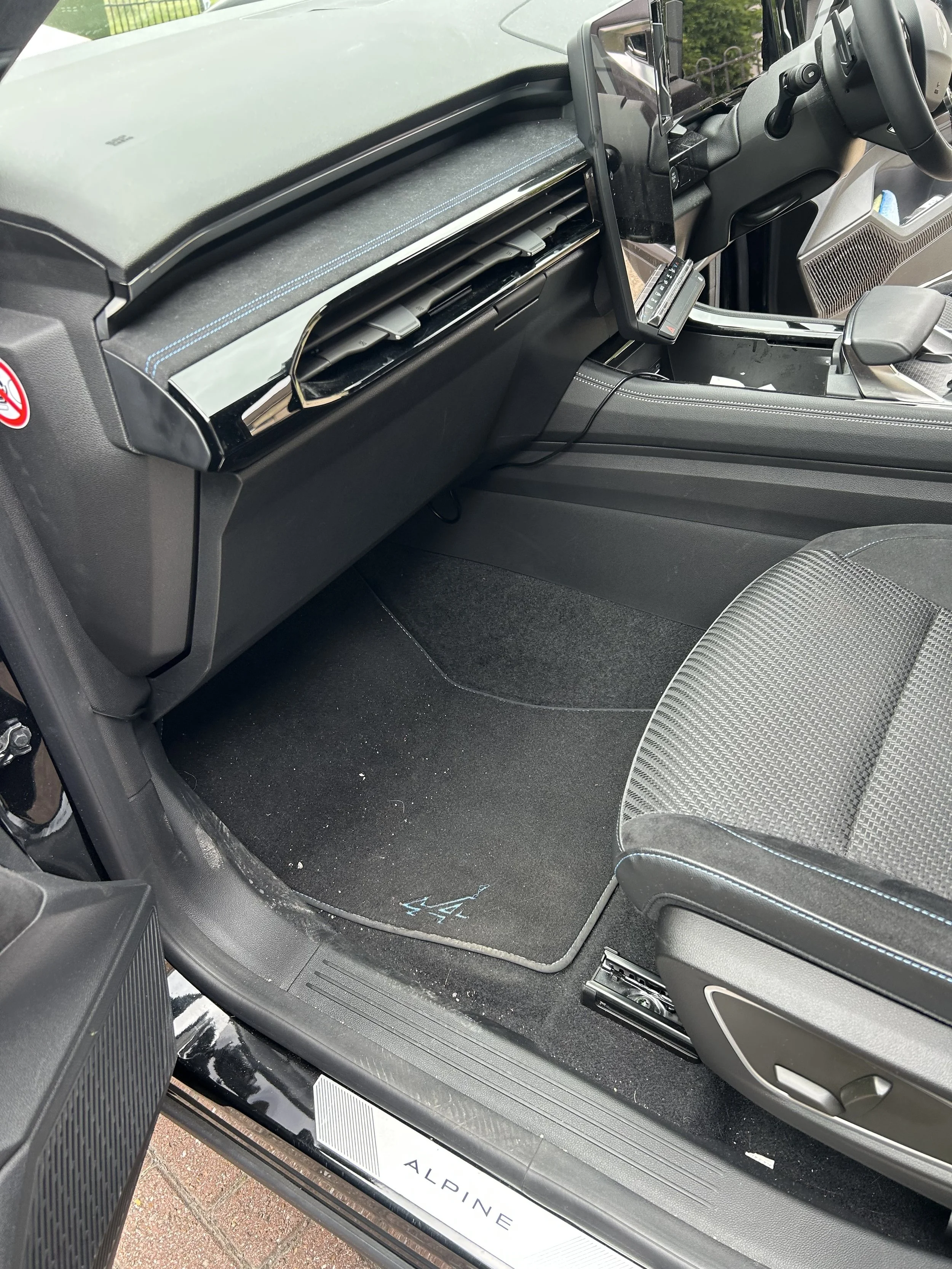 Interior of a vehicle showing the passenger side floor area, with a black floor mat, a seat, and dashboard controls. The dashboard has a mounted tablet or screen, and an air vent is visible.
