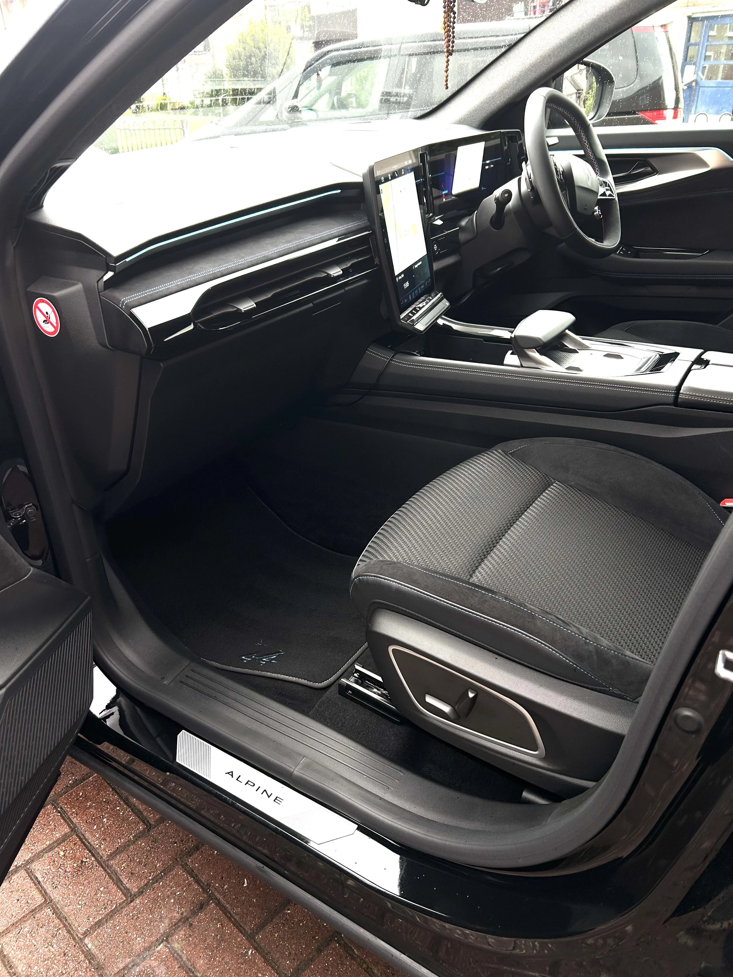 The interior of a modern vehicle, specifically a car's passenger side with a focus on the dashboard, steering wheel, and center console. The dashboard features a wide central display screen, and the interior is predominantly black with contrasting st