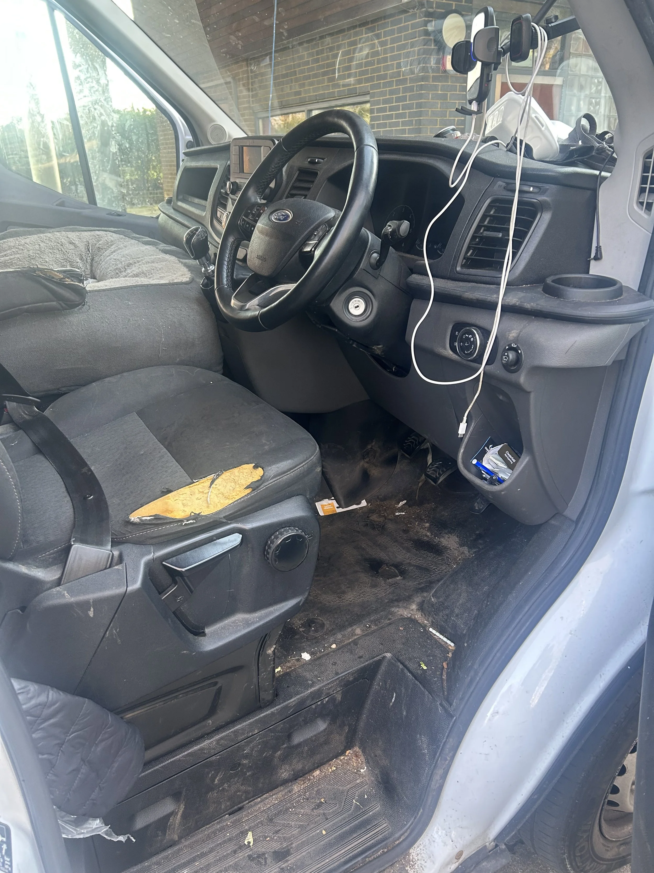 Inside a vehicle, showing the driver's seat and dashboard with visible wear and damage, including a torn seat cover. The floor area appears dirty with debris. Various wires and electronic accessories are attached to the dashboard and hanging inside t