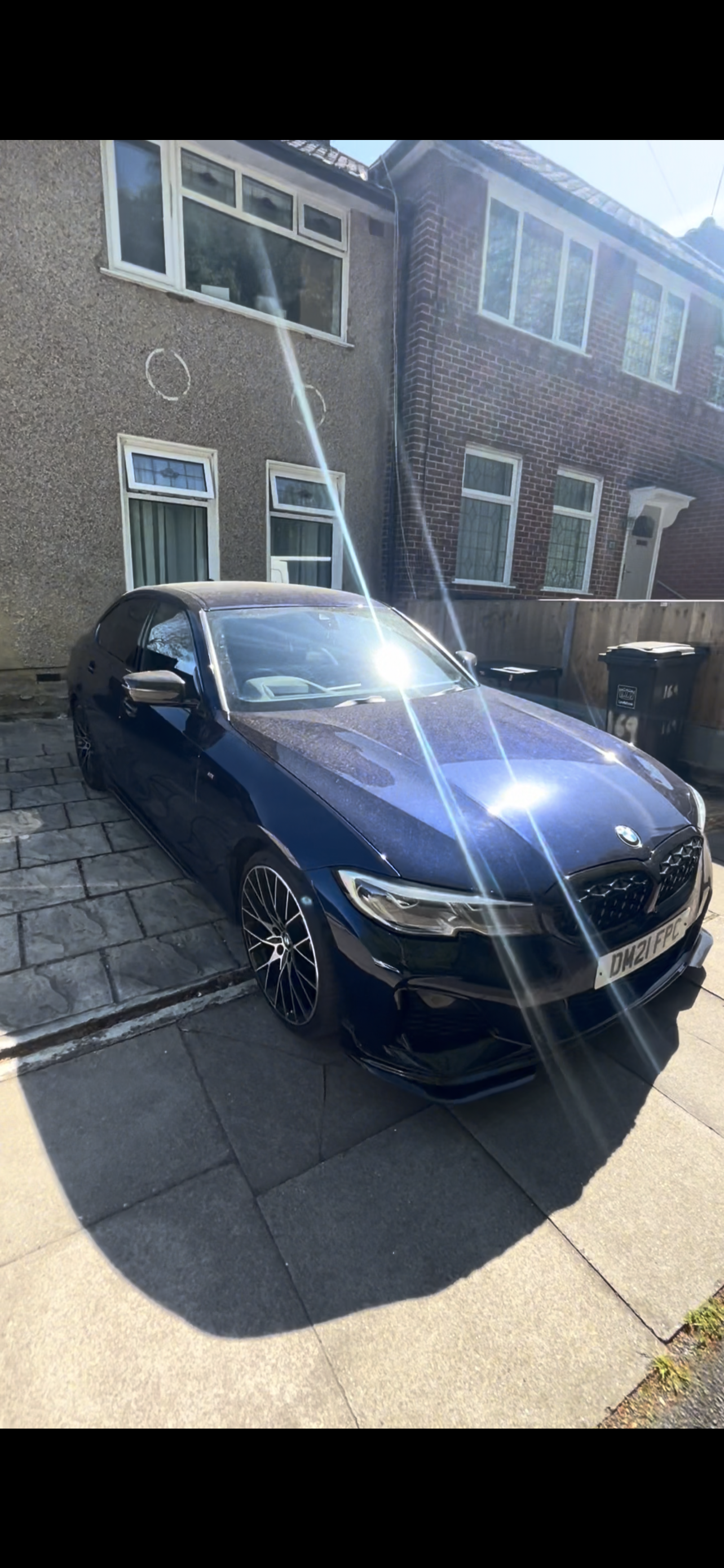 A dark blue BMW parked on a paved driveway in front of a residential house with brick and pebble-dash walls, with sunlight creating lens flares in the image.