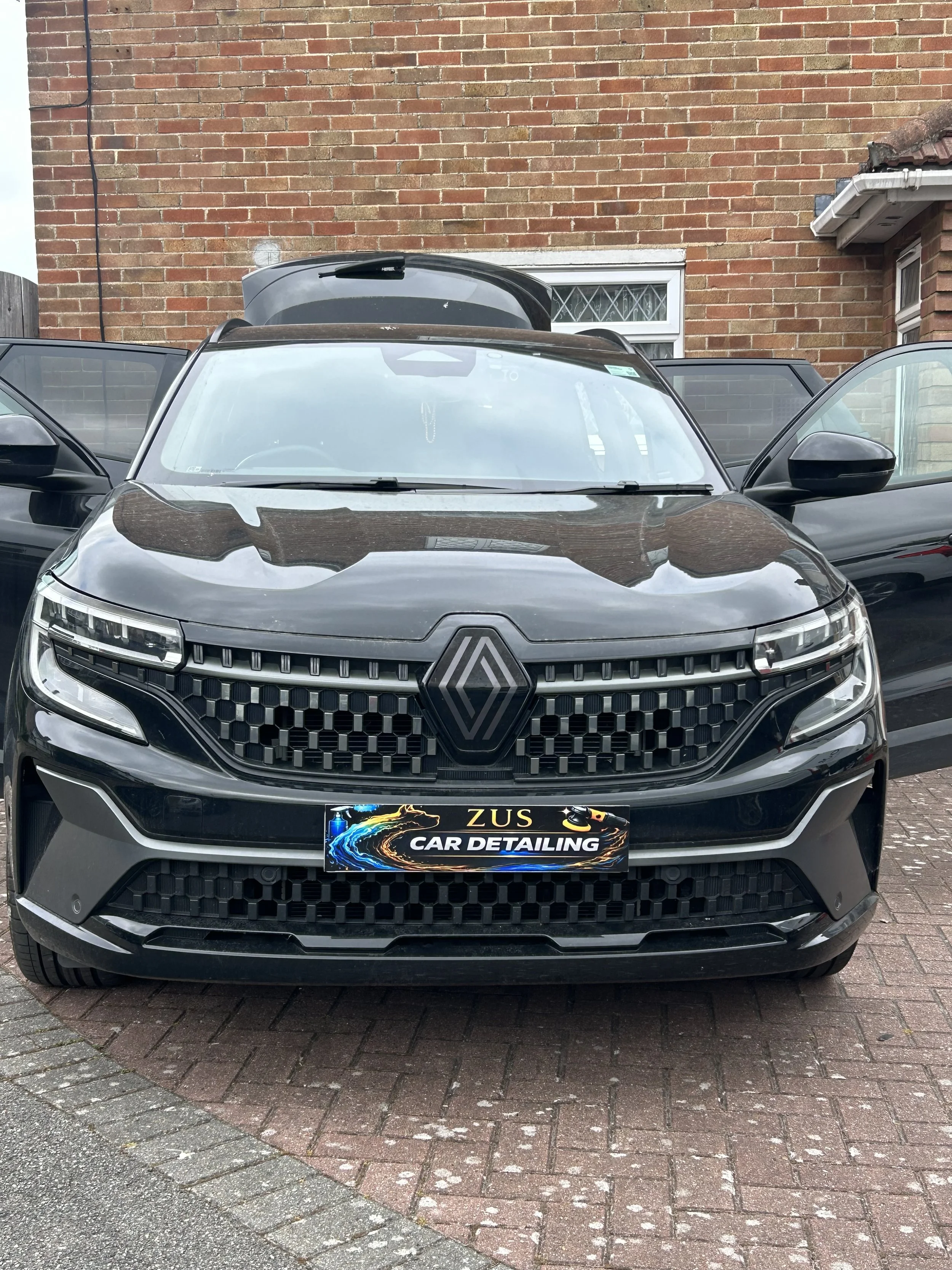 Front view of a black Renault car parked on a brick driveway with open doors, a brick wall in the background, and a sign on the front bumper that reads 'ZUS CAR DETAILING'.