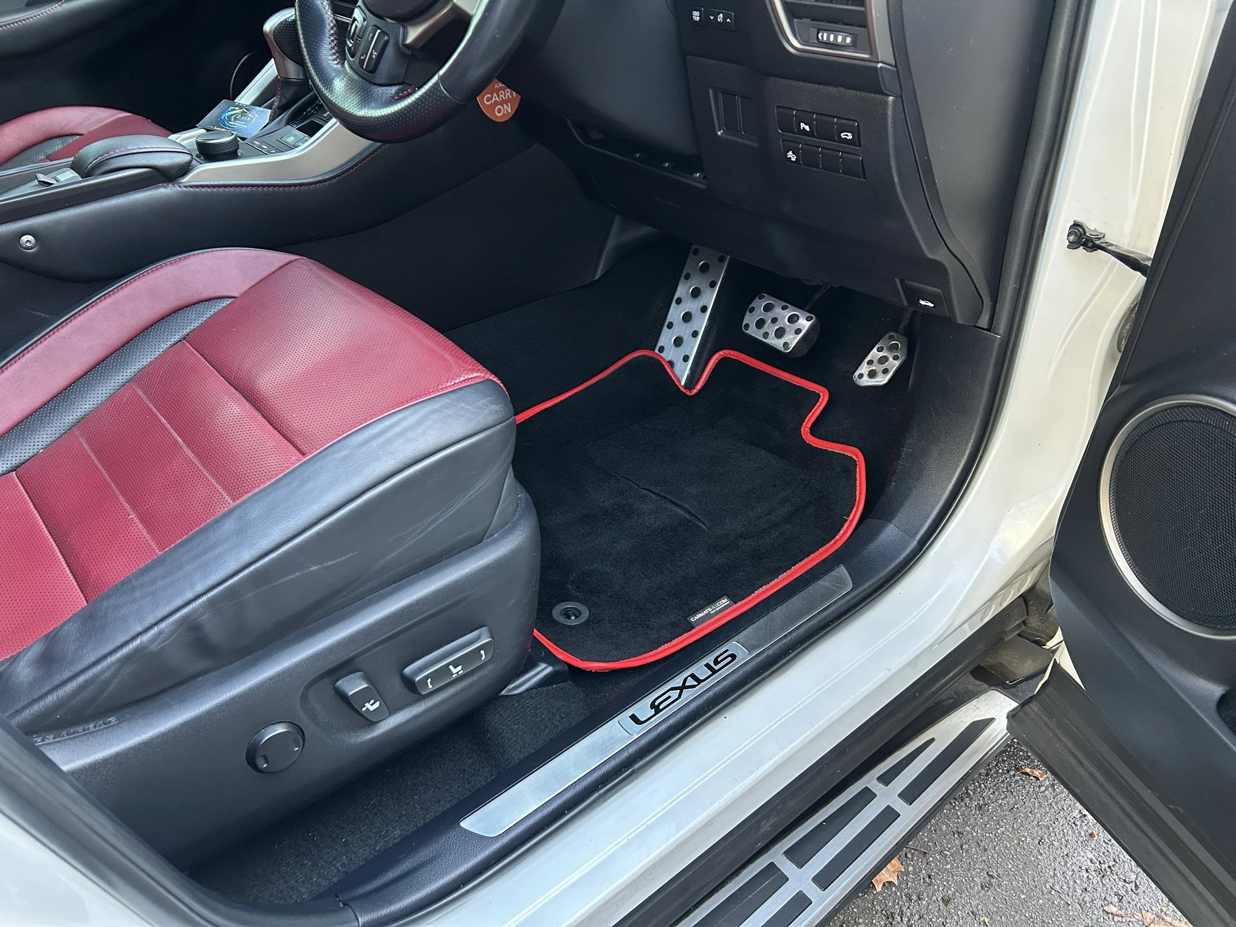 Interior of a car showing the driver's seat, pedals, and dashboard. The seat is black and red, and the pedals are metallic with holes. There is a red and black mat on the floor with red stitching. The door sill has a silver trim with the word 'LEXUS'