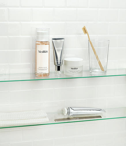 Medik8 skincare products, a toothbrush in a glass, and other personal care items on glass shelves in a bathroom with white subway tiles.