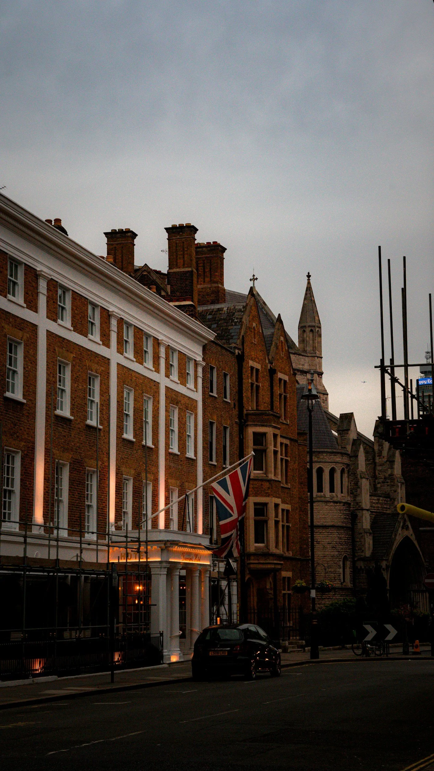 City street with historic brick buildings, church steeple, and Union Jack flag, under gray overcast sky, parked cars, and scaffolding.