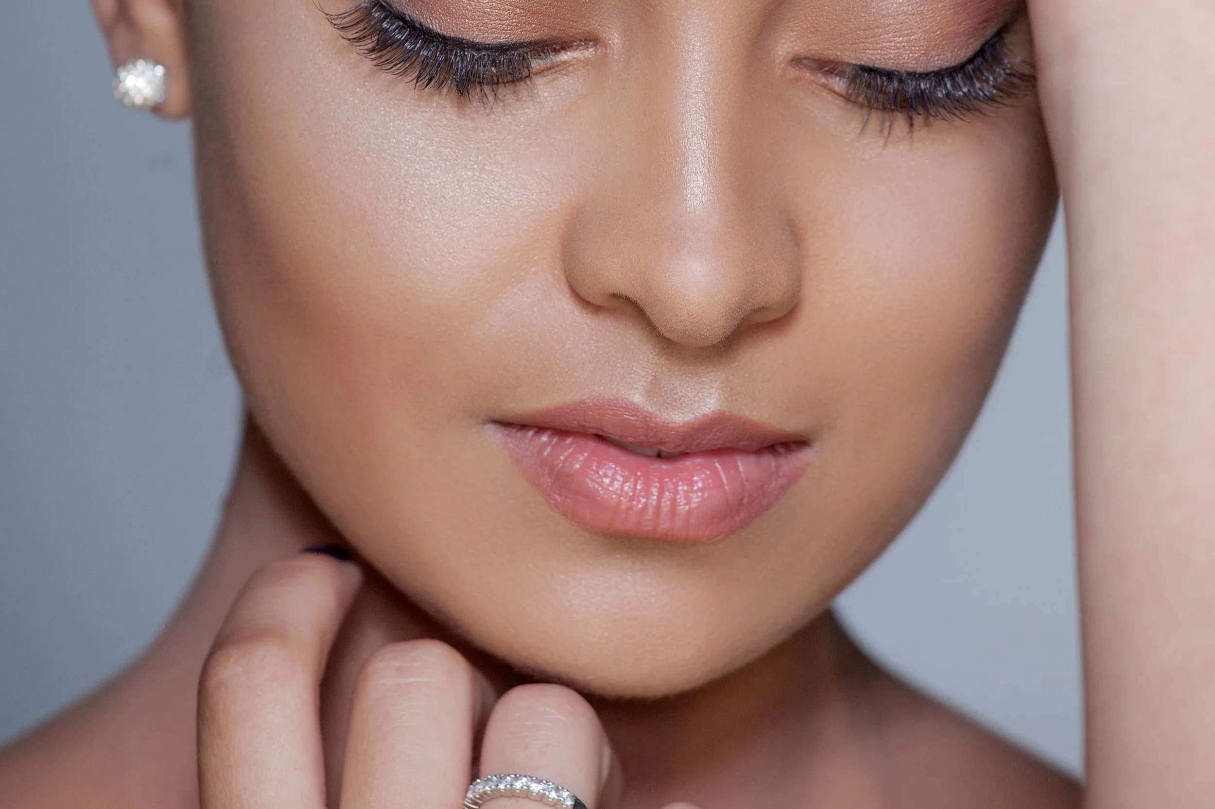 Close-up of a woman with closed eyes, wearing earrings and a ring, touching her face gently.