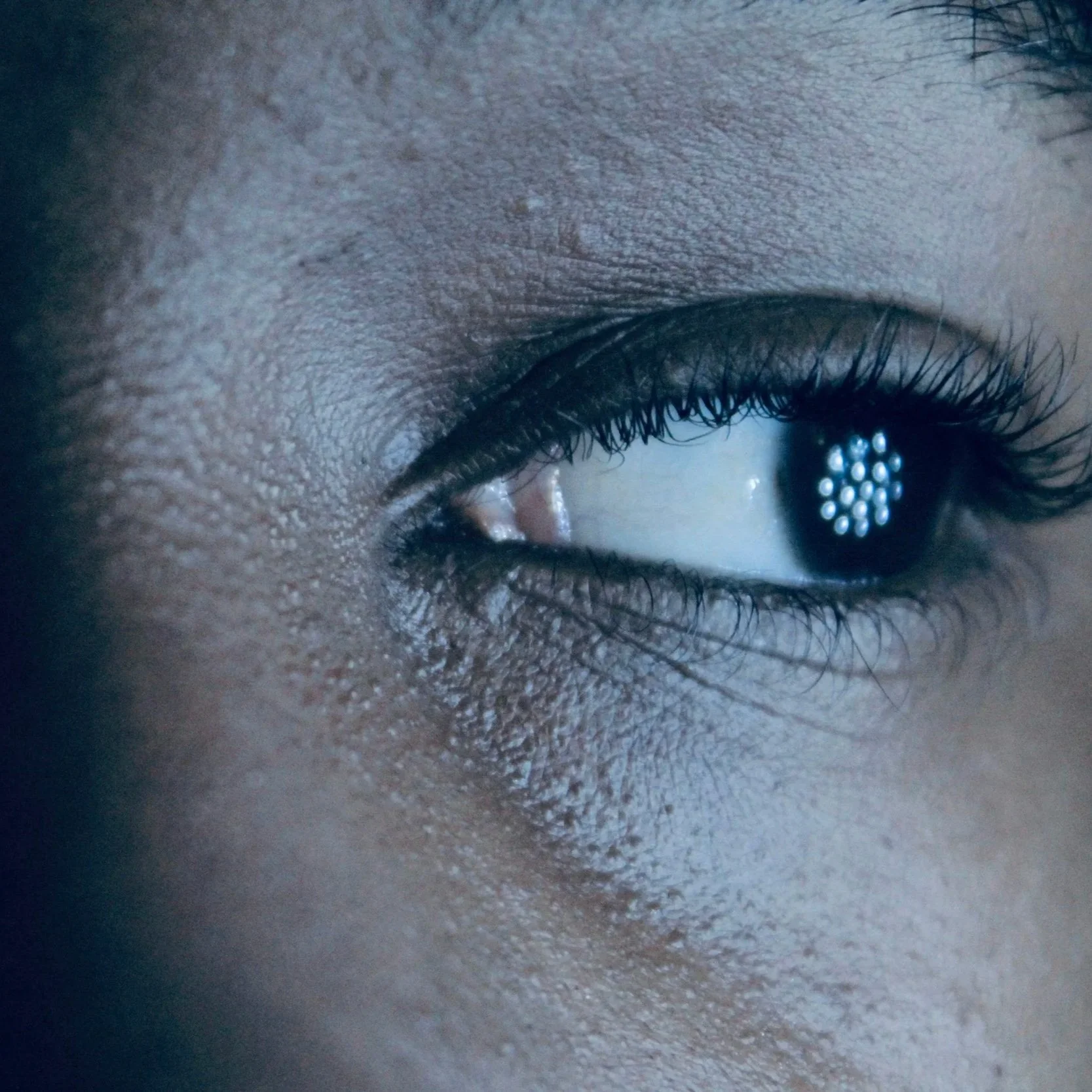 Close-up of a person's eye with long eyelashes, part of the eyebrow and the skin around the eye showing texture and fine details.