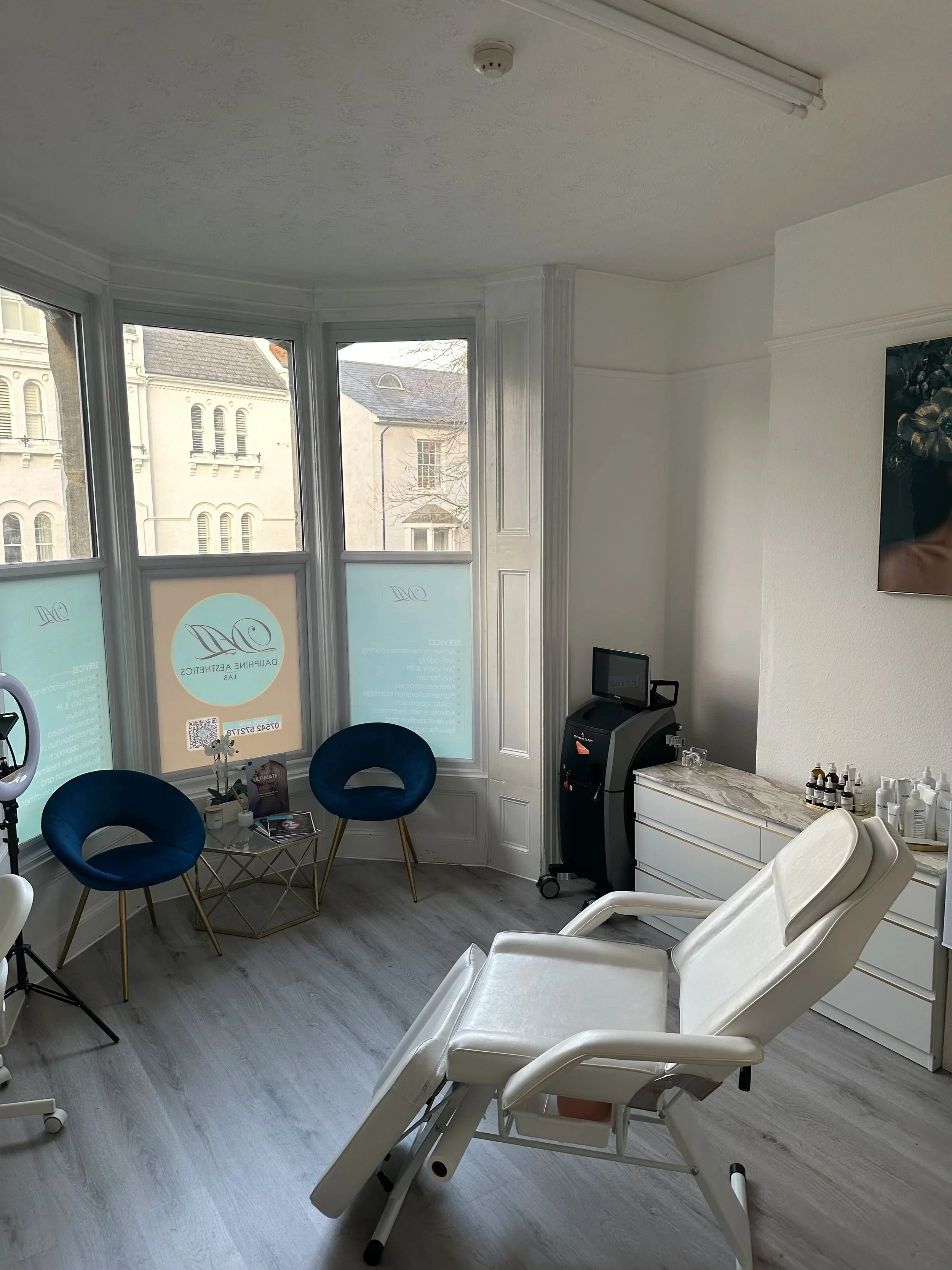 Interior of a medical or spa clinic room with a white treatment chair, two blue chairs with gold legs, a table, and medical supplies on a white cabinet. Large windows let in natural light, revealing historic buildings outside.DAL Swindon