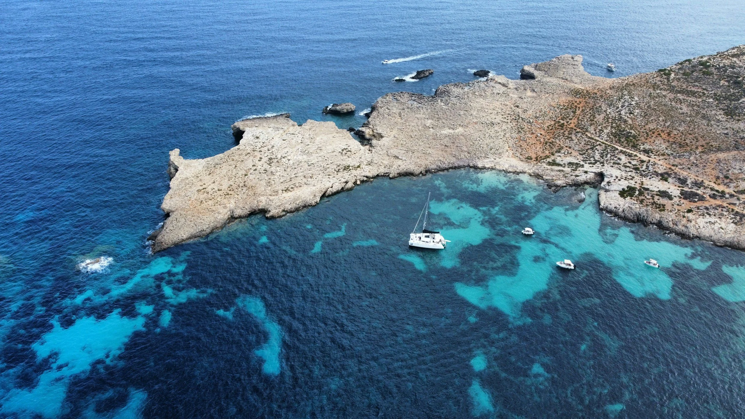 An aerial view of a rocky coastline with a small bay featuring a white sailboat and several smaller boats, surrounded by clear blue water.