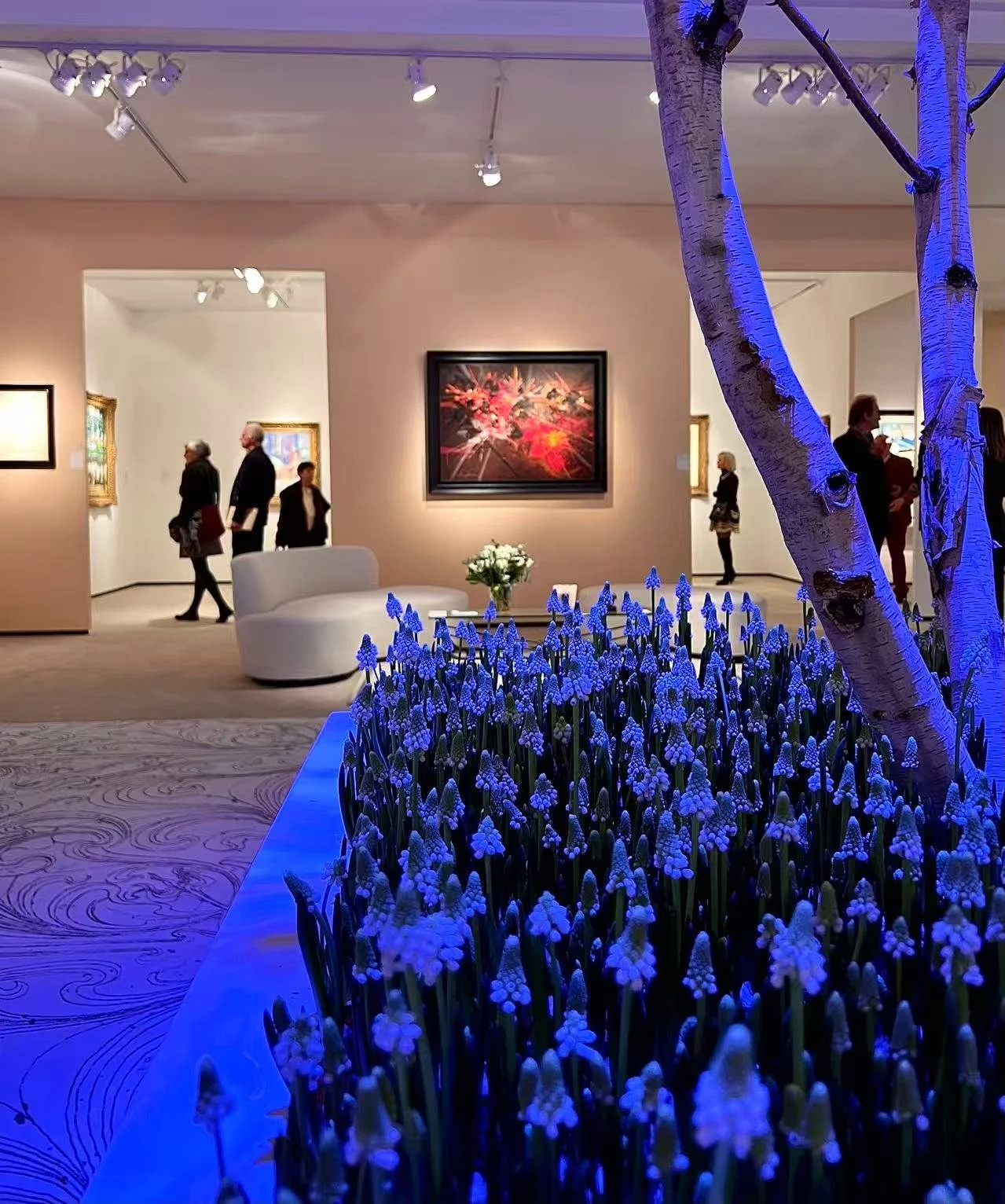 An art gallery with people viewing artwork. In the foreground, there are blue flowers and a tree with white bark illuminated by blue lighting. The gallery walls display framed paintings, and there is a white sofa with a small flower arrangement on the table nearby.