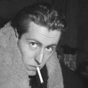 Black and white photo of a man with slicked-back hair smoking a cigarette, wearing a coat.