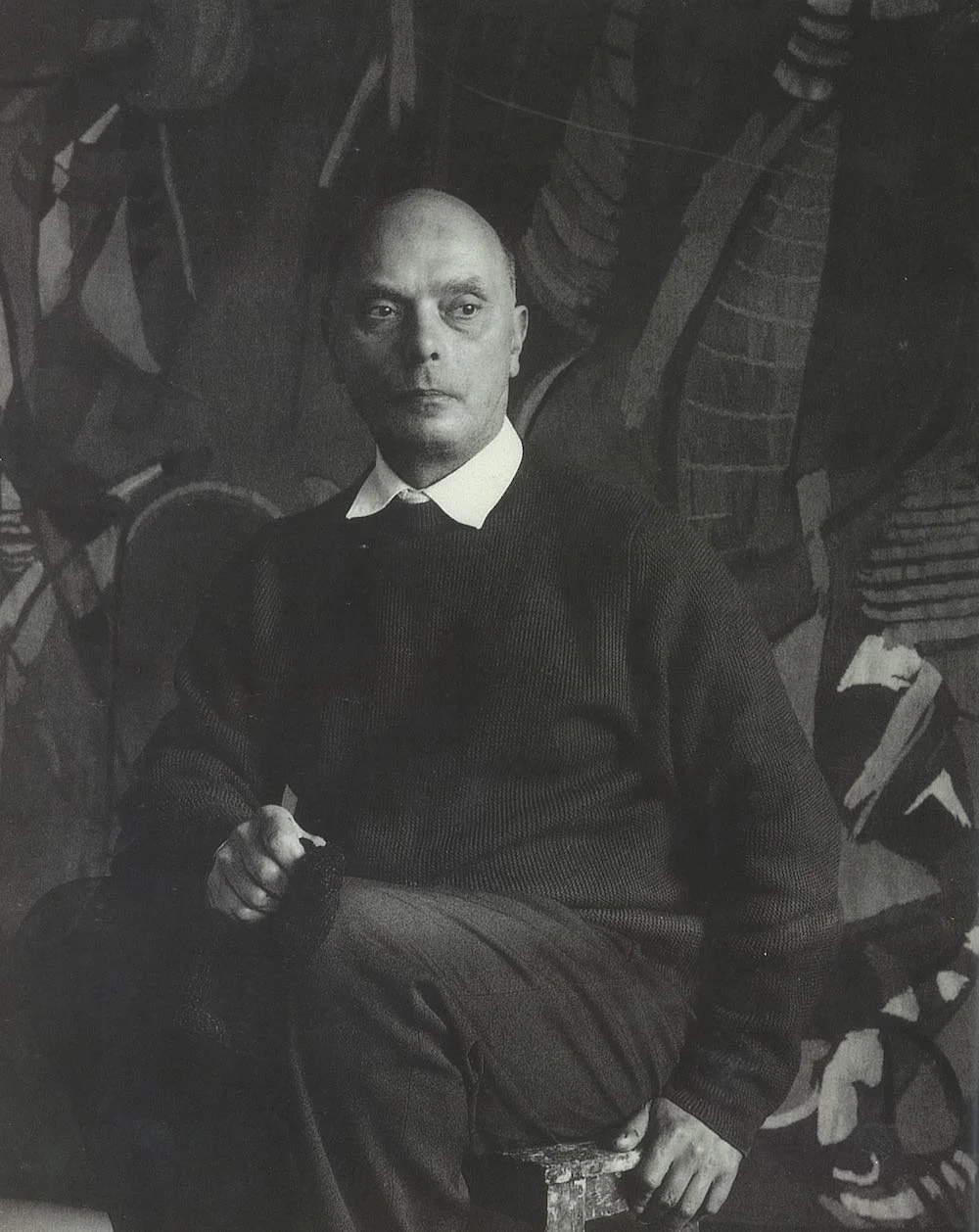 Black and white photograph of a man with a serious expression, seated in front of an abstract mural. He is wearing a dark sweater over a white collared shirt.
