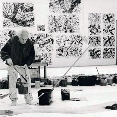 An elderly man with white hair working on an abstract painting in an art studio, surrounded by various black and white artworks on the wall.