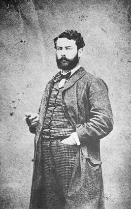 A young man with a beard and dark curly hair, wearing a coat and patterned trousers, standing with one hand in his pocket and the other holding a small object, in front of a plain background.