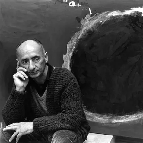 Man sitting with his chin resting on his hand, next to a large, dark circular abstract painting on a wall.