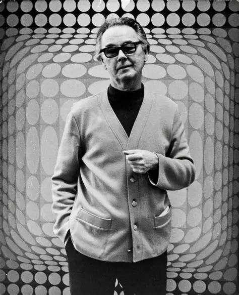 A man wearing glasses and a light cardigan over a dark turtleneck, standing in front of a retro geometric patterned background.