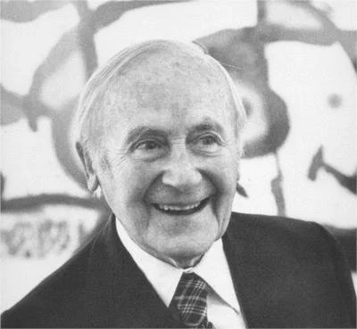 An elderly man with white hair smiling, wearing a suit jacket and tie, in front of a blurred artistic background.
