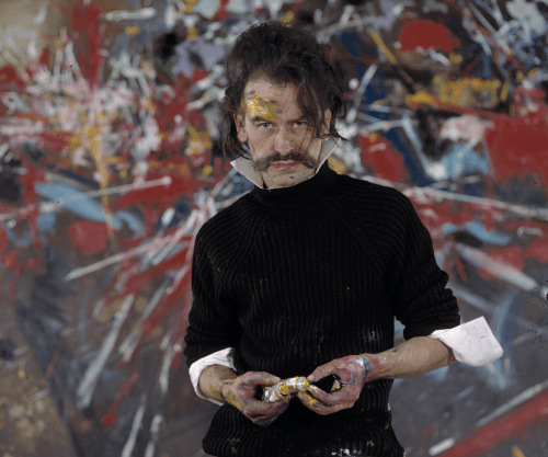 A man with dark, disheveled hair, mustache, and paint on his face and hands, wearing a black sweater with rolled sleeves, standing in front of a colorful, abstract painted background.