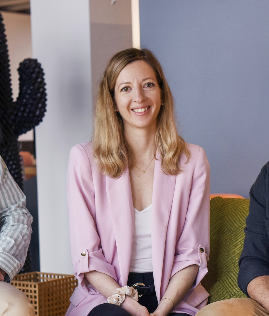Aude, Freelance en paid media en Suisse, Allemagne et France, assise, souriant, portant une veste rose et un t-shirt blanc, dans un intérieur moderne.