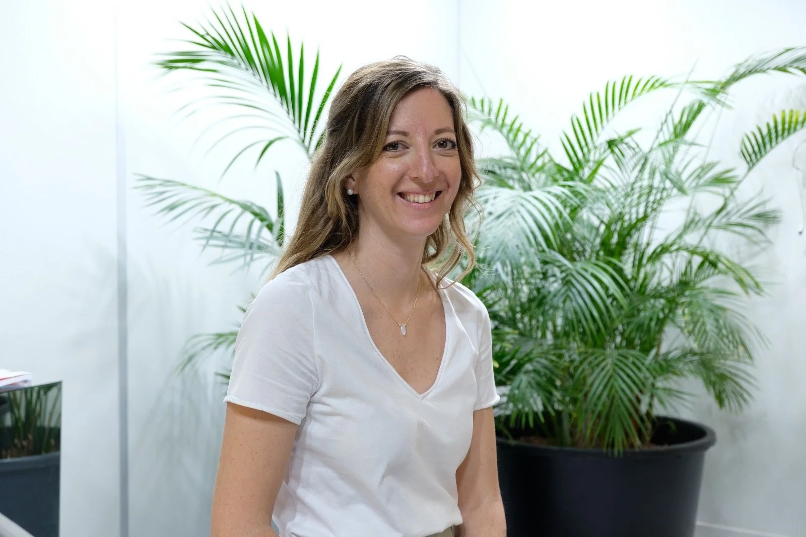 Aude, Freelance en paid media en Suisse, Allemagne et France, souriante portant un t-shirt blanc, posé devant un grand pot de plantes vertes d'intérieur.
