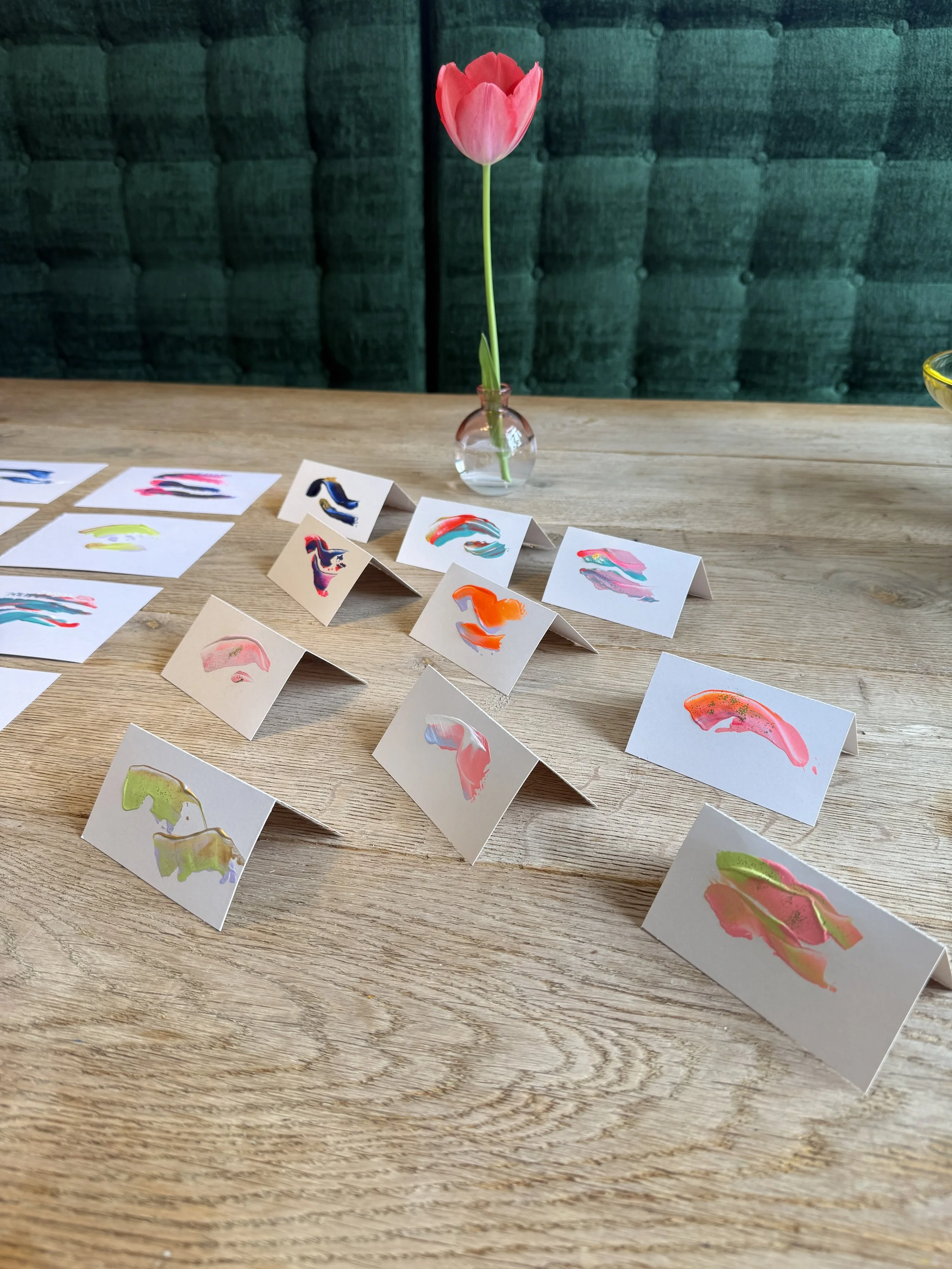 A pink tulip in a small round glass vase on a wooden table, with numerous colorful abstract art pieces on folded paper cards arranged around it.