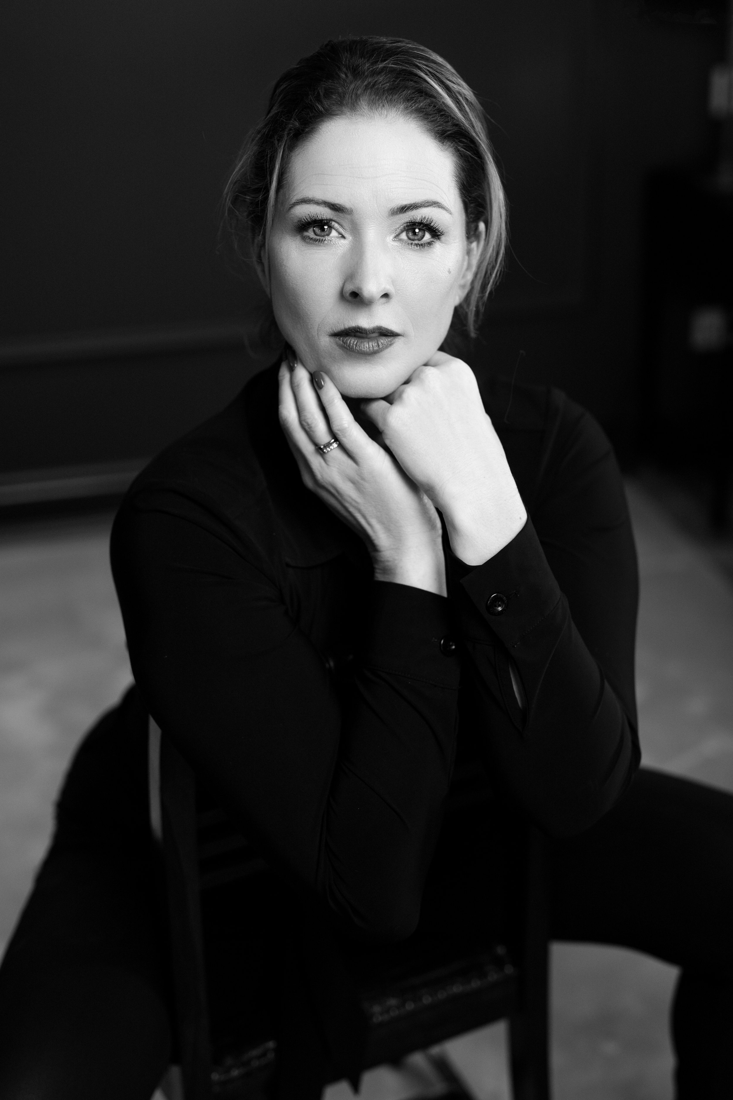 A black-and-white portrait of a woman with long hair, sitting on a chair, wearing a dark top, with her hands gently resting on her chin, looking directly at the camera.