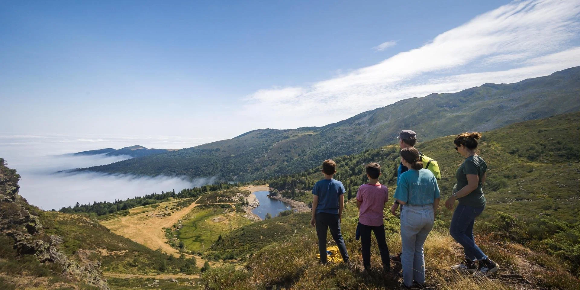 Groupe de personnes en randonnée dans un paysage de montagne avec une rivière, des collines verdoyantes, des nuages en basse altitude et un ciel bleu clair.