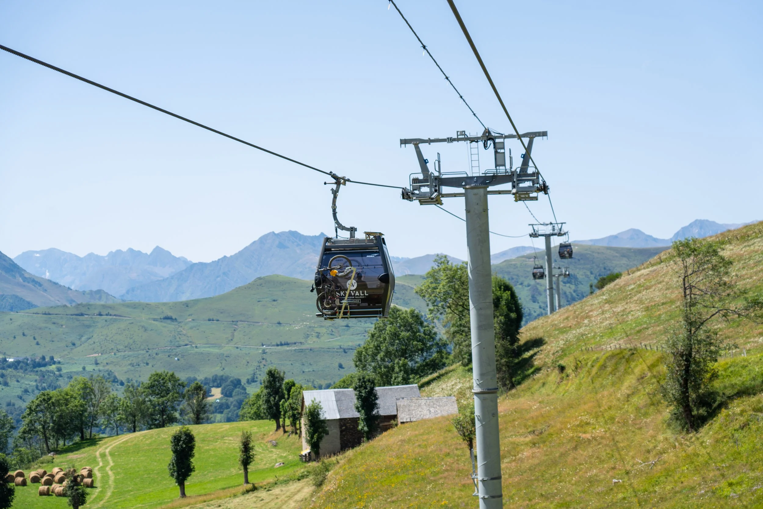 Téléphérique avec des cabines suspendues, traversant un paysage rural avec des collines verdoyantes, quelques maisons et des montagnes en arrière-plan
