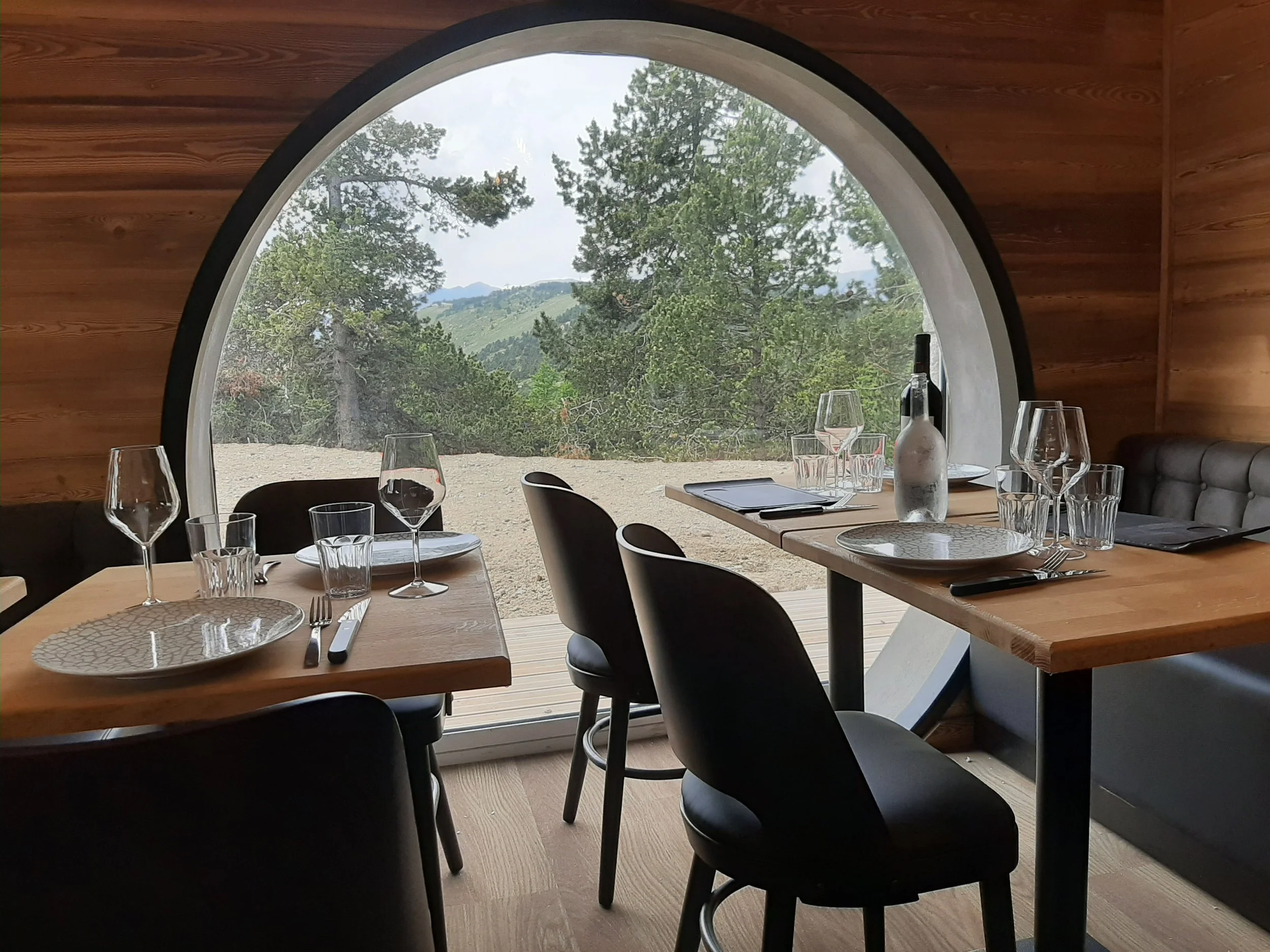 Vue intérieure d'une salle à manger avec une grande fenêtre de forme arrondie, affichant une vue de montagnes et d'arbres. La table est dressée avec des verres, assiettes, une bouteille de vin, et une tablette ou fiche informatique. Le mobilier est en bois avec des chaises noires, et l'intérieur est en bois naturel.