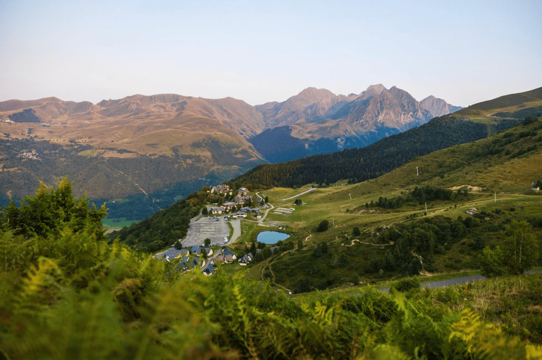 Paysage montagneux avec des collines verdoyantes, une petite ville et un lac, sous un ciel clair.