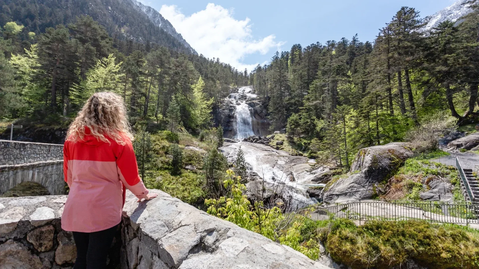 Une personne avec les cheveux bouclés portant une veste rouge admire une cascade au milieu d'une forêt verdoyante montagnarde.