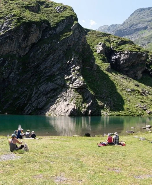 Groupe de personnes assises près d'un lac entouré de montagnes rocheuses et verdoyantes, par une journée ensoleillée.
