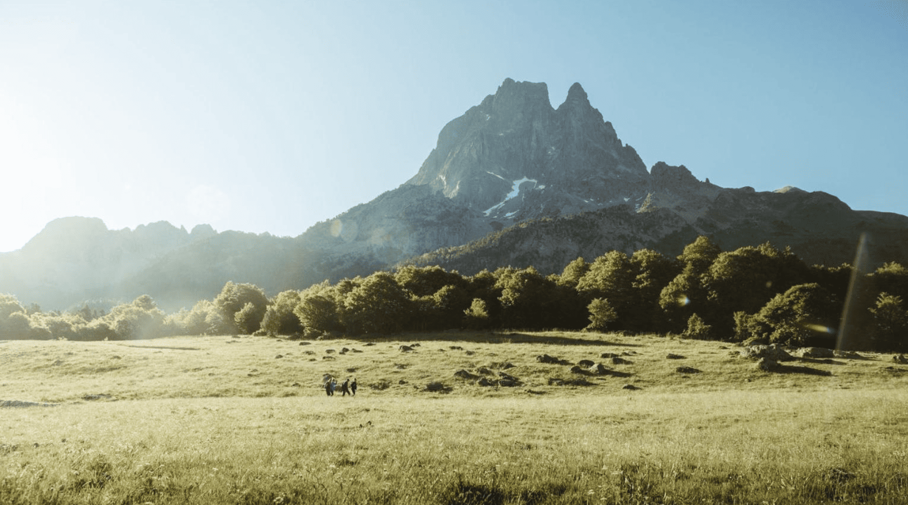 Paysage de montagne avec sommet rocheux, forêt dense à la base et prairie en avant avec deux personnes marchant.