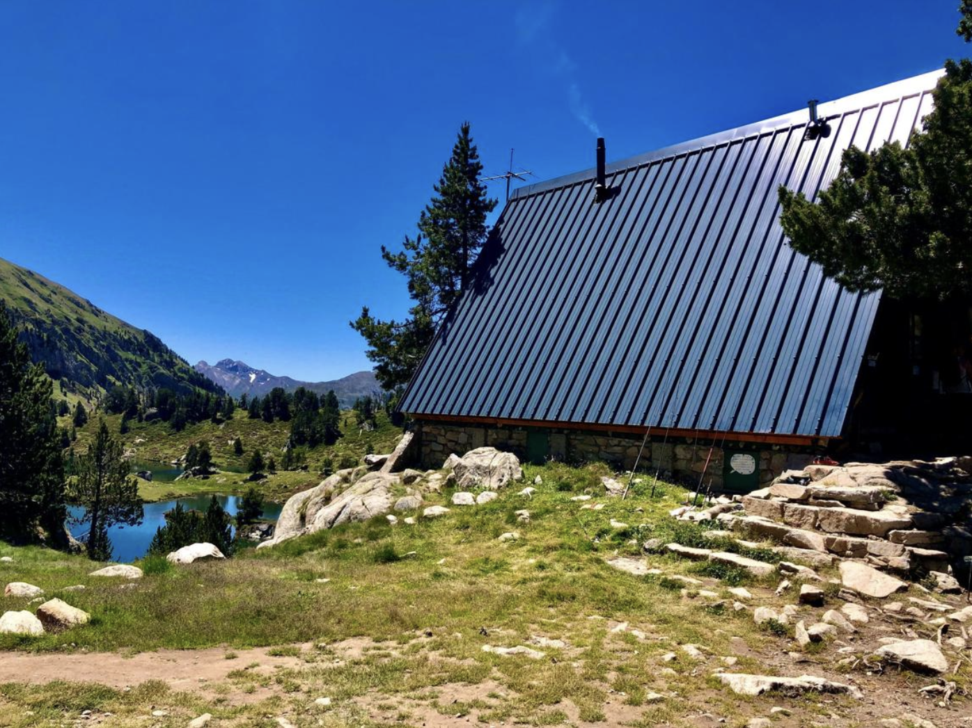 Cabane en pierre avec un toit en métal bleu située en montagne, entourée d'arbres, avec lac et montagnes en toile de fond sous un ciel bleu clair.