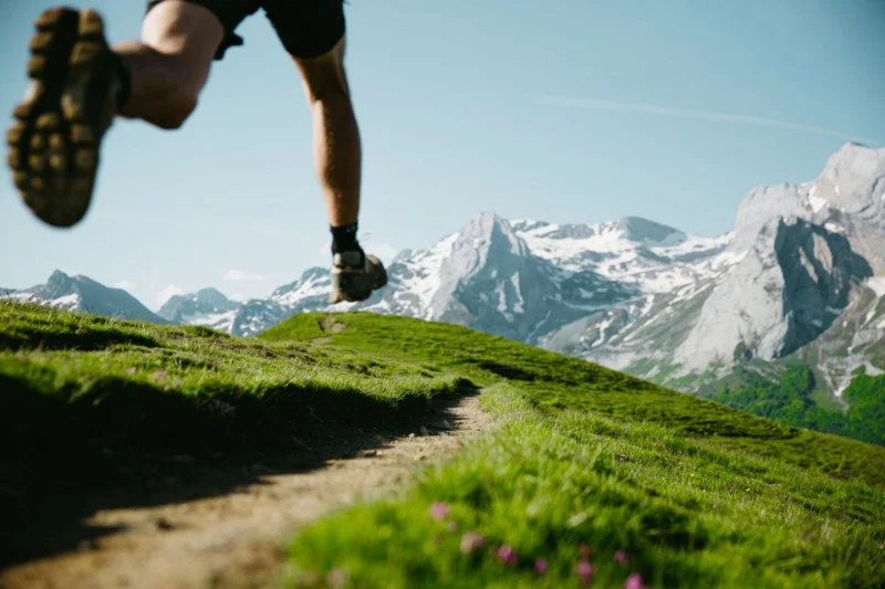 Un coureur en pleine course sur un sentier de montagne avec des sommets enneigés en arrière-plan.