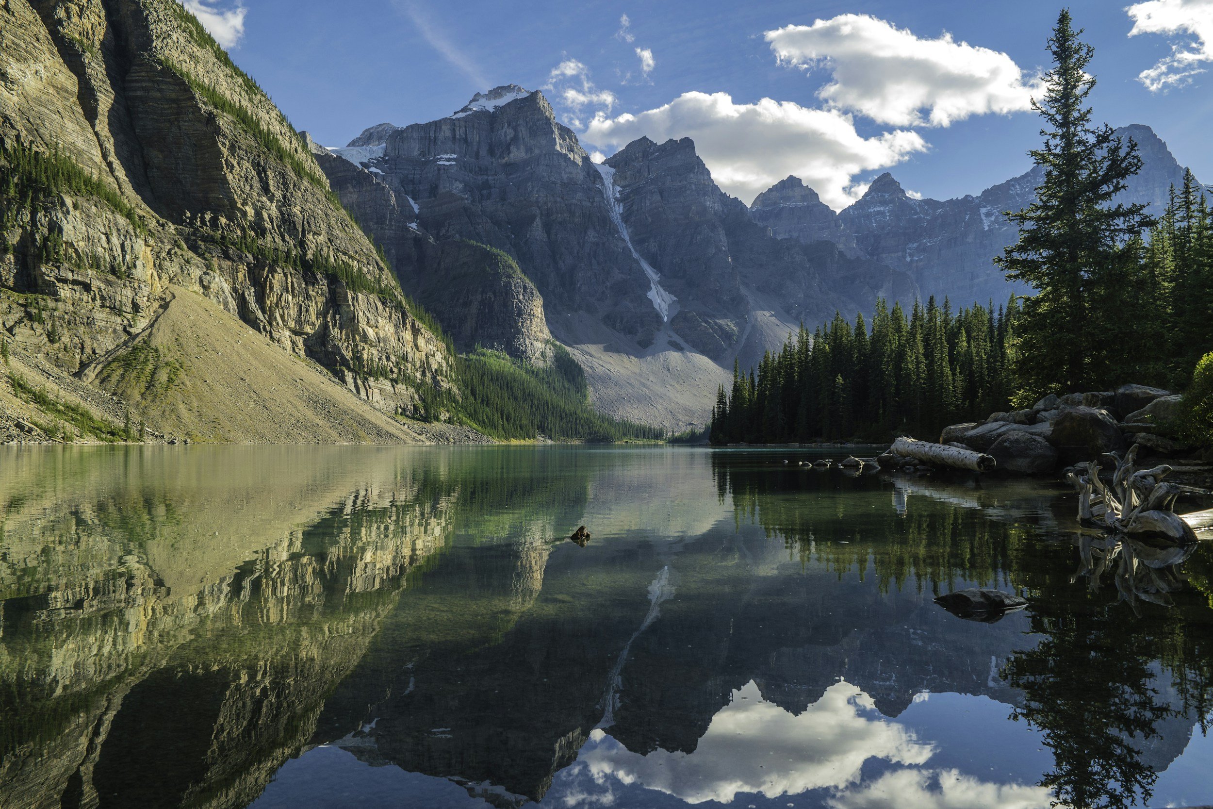 Un lac calme entouré de montagnes rocheuses avec des sommets enneigés et un ciel bleu avec quelques nuages, reflet parfait dans l'eau.