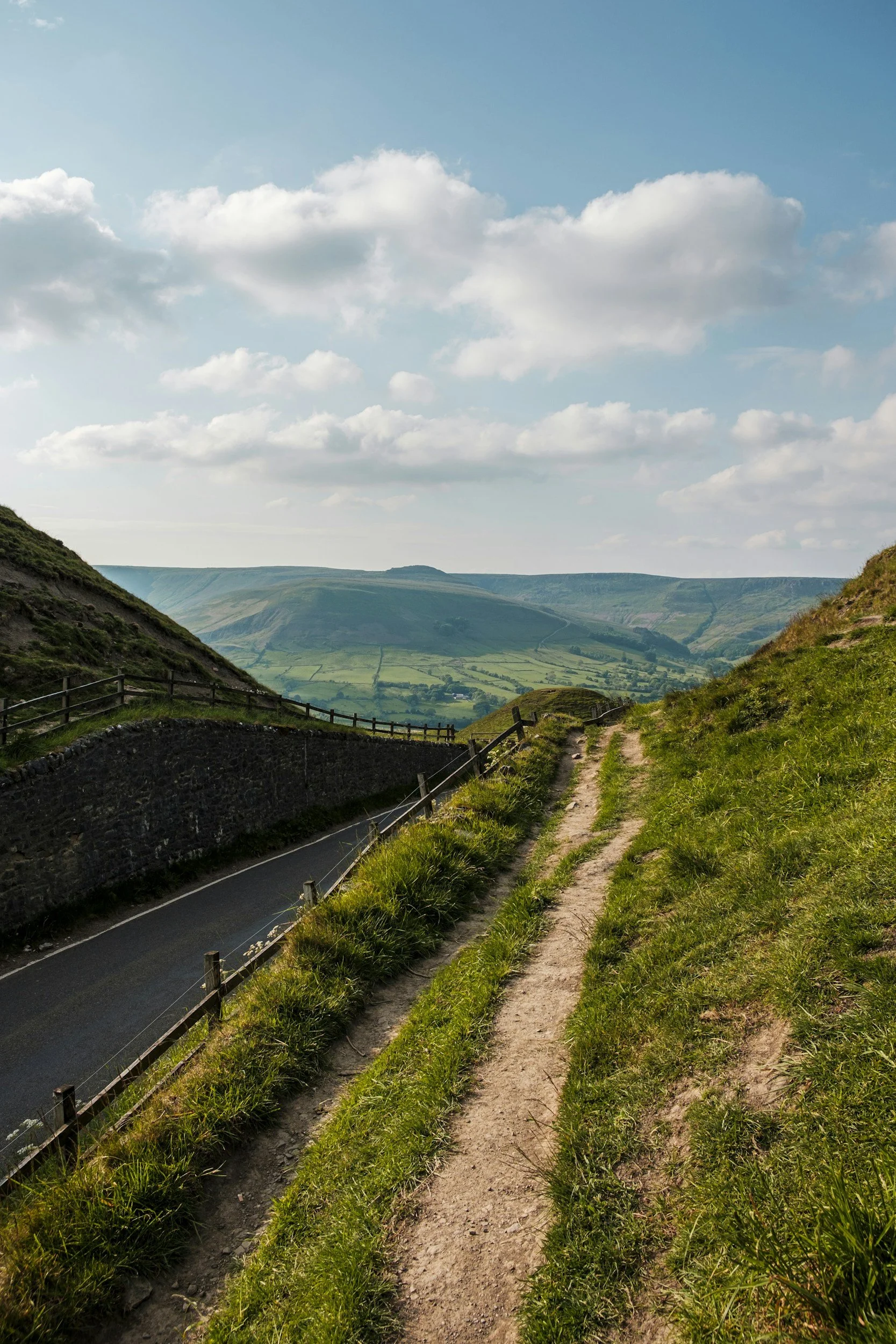 Chemin de terre sur une colline verte surplombant une vallée avec des collines en arrière-plan, sous un ciel bleu avec quelques nuages.