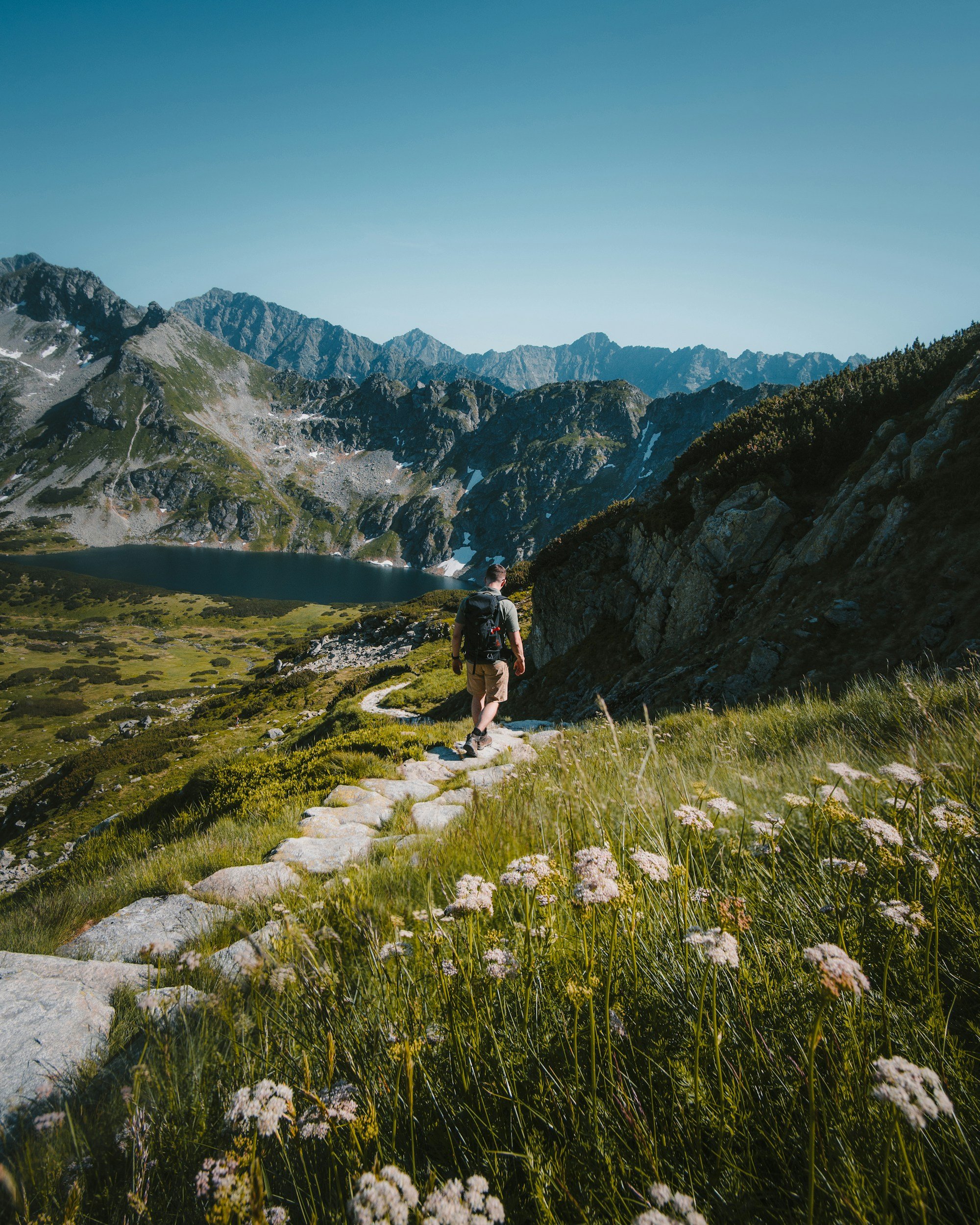 Une personne avec un sac à dos marche sur un sentier de pierre dans une vallée alpine entourée de montagnes avec un lac en contrebas.