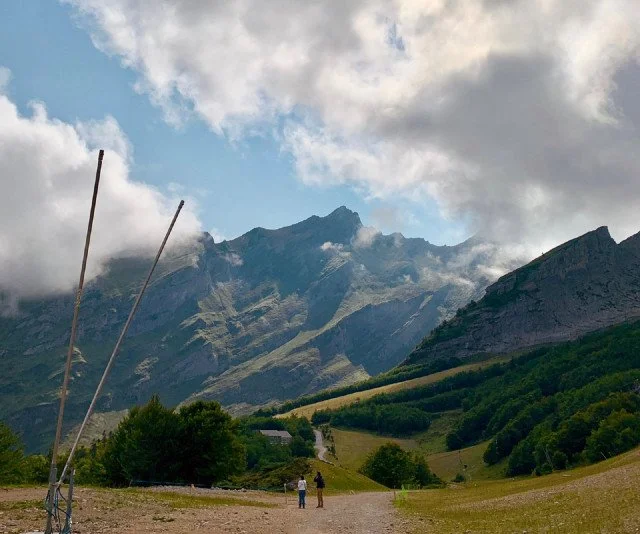 Deux personnes marchant dans une vallée verdoyante avec des montagnes en arrière-plan et un ciel nuageux.