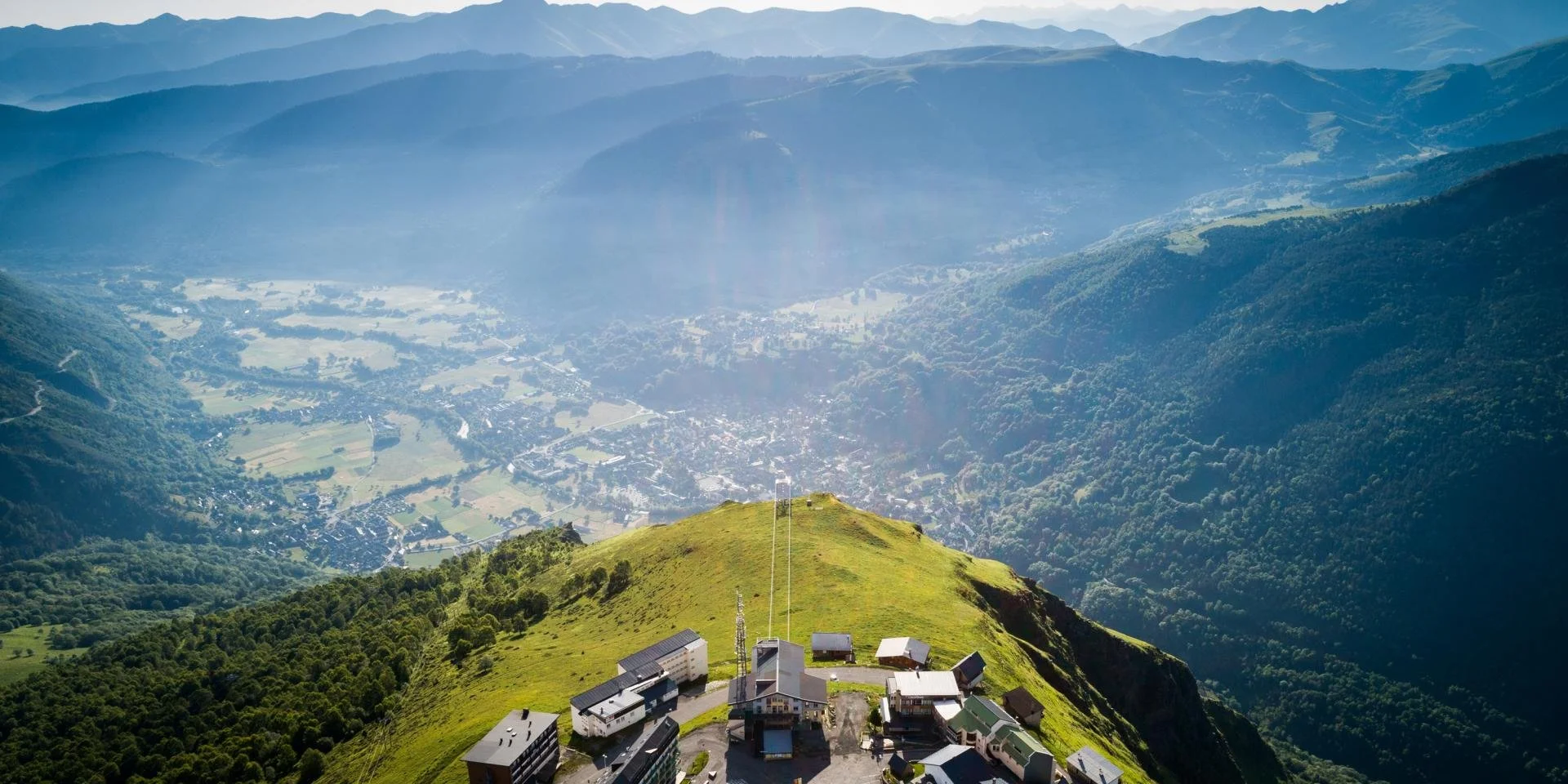 Photo d'une station de montagne en hauteur, avec bâtiments, entourée de verdure, et une vue panoramique de la vallée en contrebas avec des montagnes en arrière-plan.