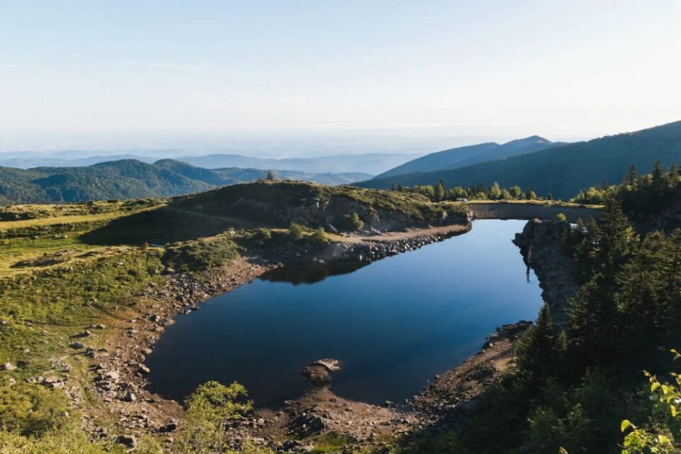 Un lac de montagne entouré de collines verdoyantes, avec des montagnes au loin sous un ciel clair.