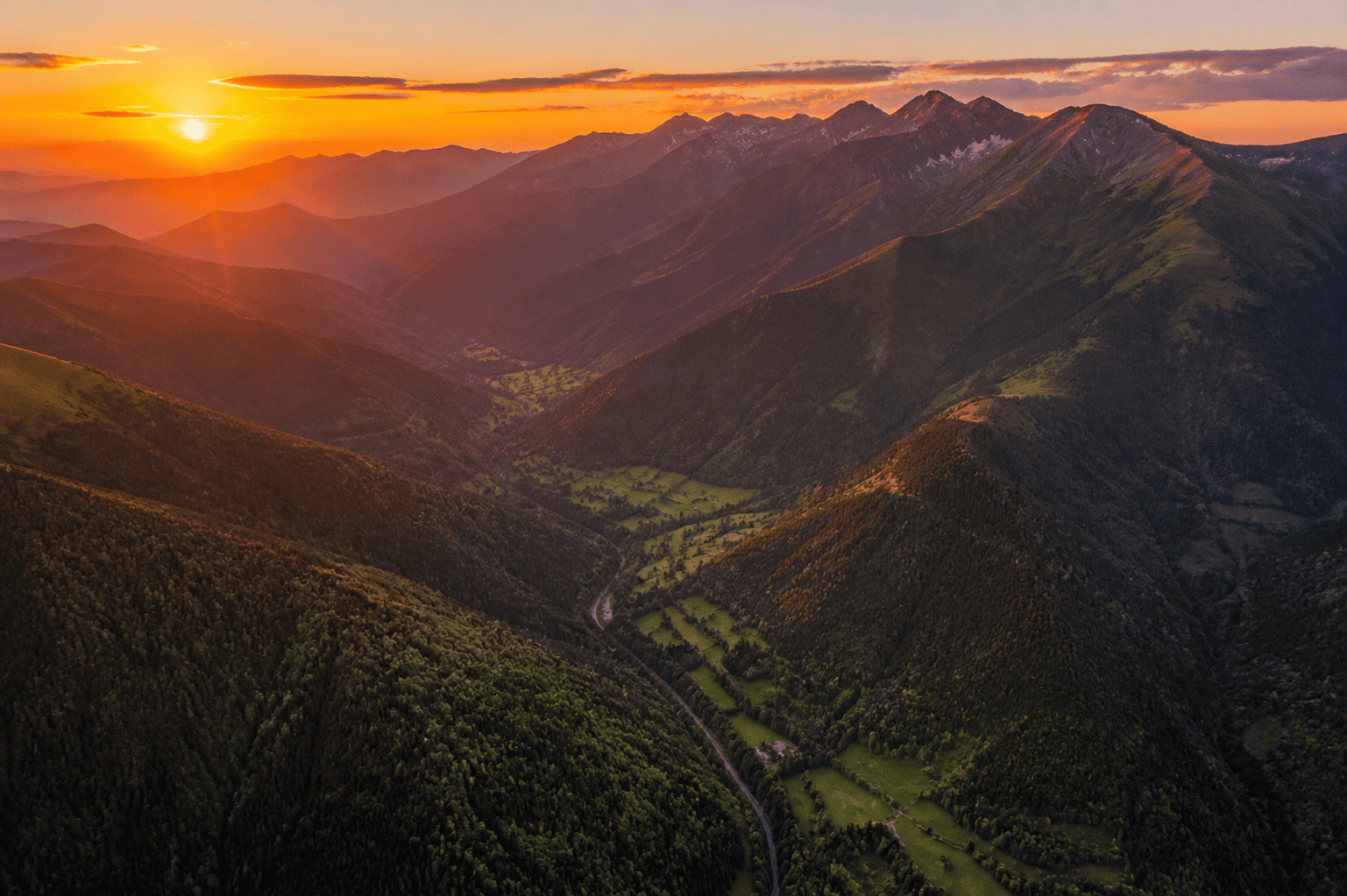 Paysage montagneux au coucher du soleil avec des vallées verdoyantes et un ciel orange.