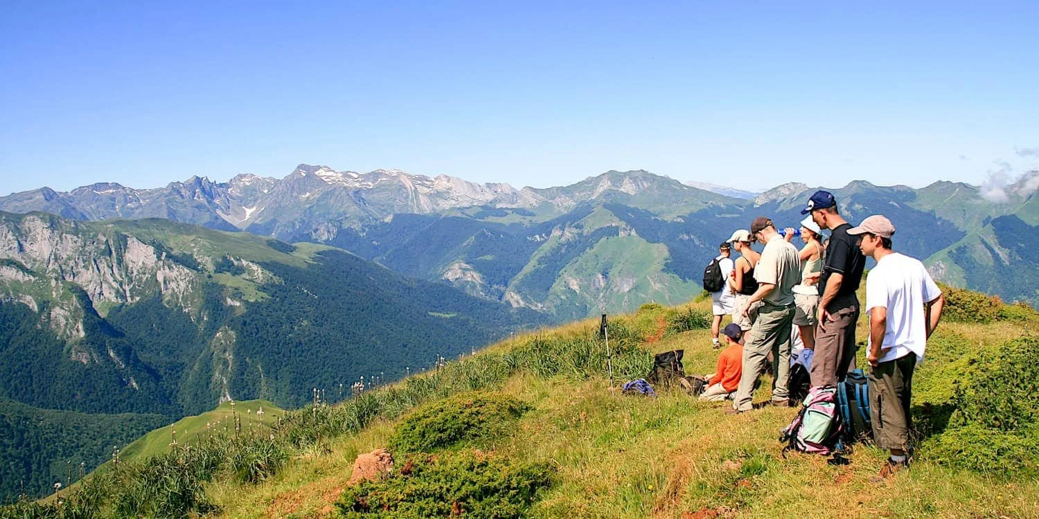 Groupe de randonneurs en pause sur une colline verdoyante avec vue sur des montagnes enneigées et des vallées en arrière-plan, sous un ciel bleu clair.