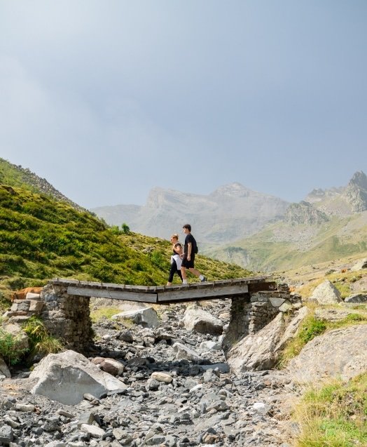 Deux personnes, un adulte et un enfant, marchent sur un petit pont en pierre dans un paysage de montagne avec des rochers et de la végétation verte.