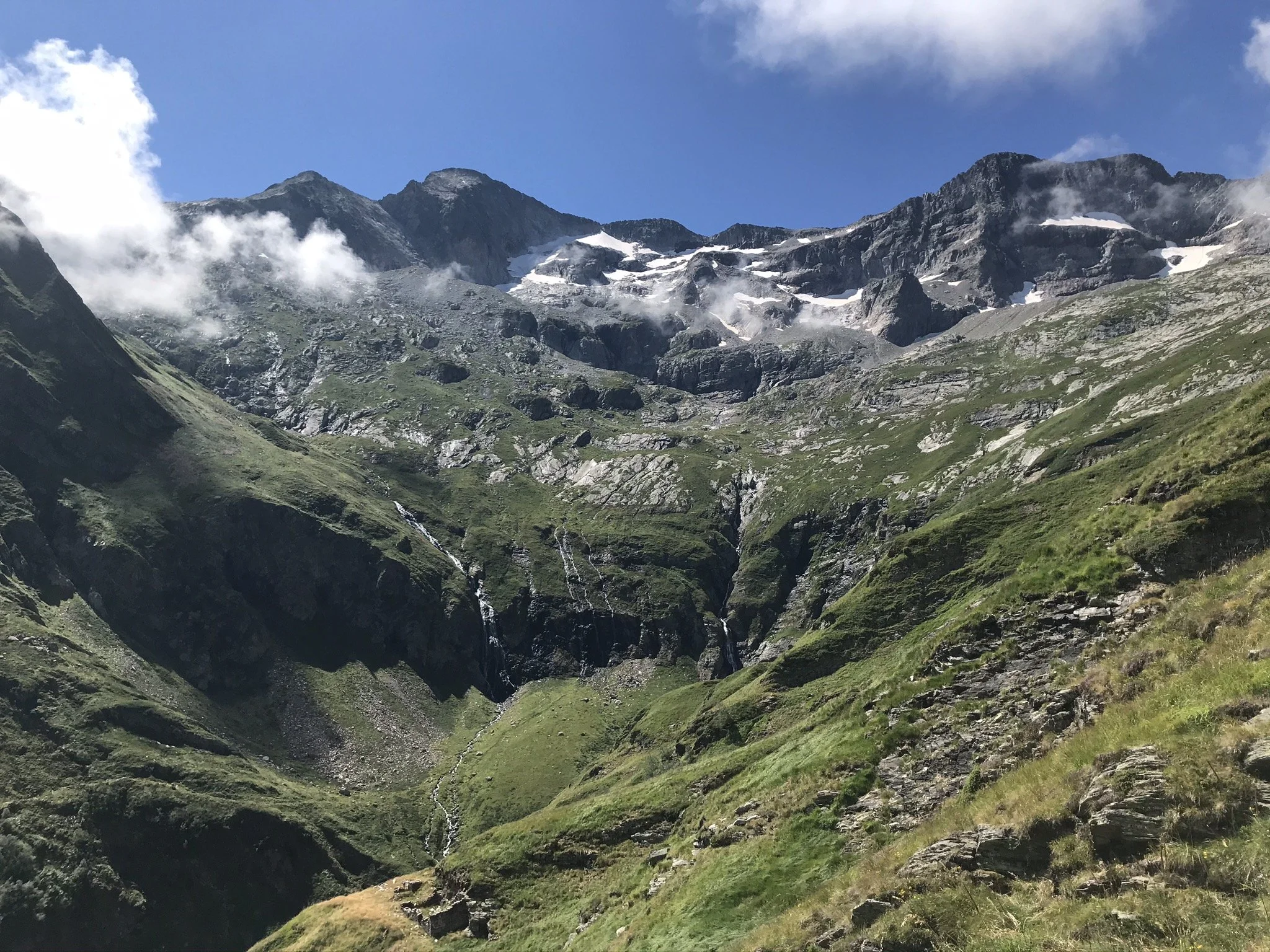 Montagnes avec des sommets enneigés, des pentes verdoyantes et des nuages dans un ciel bleu.